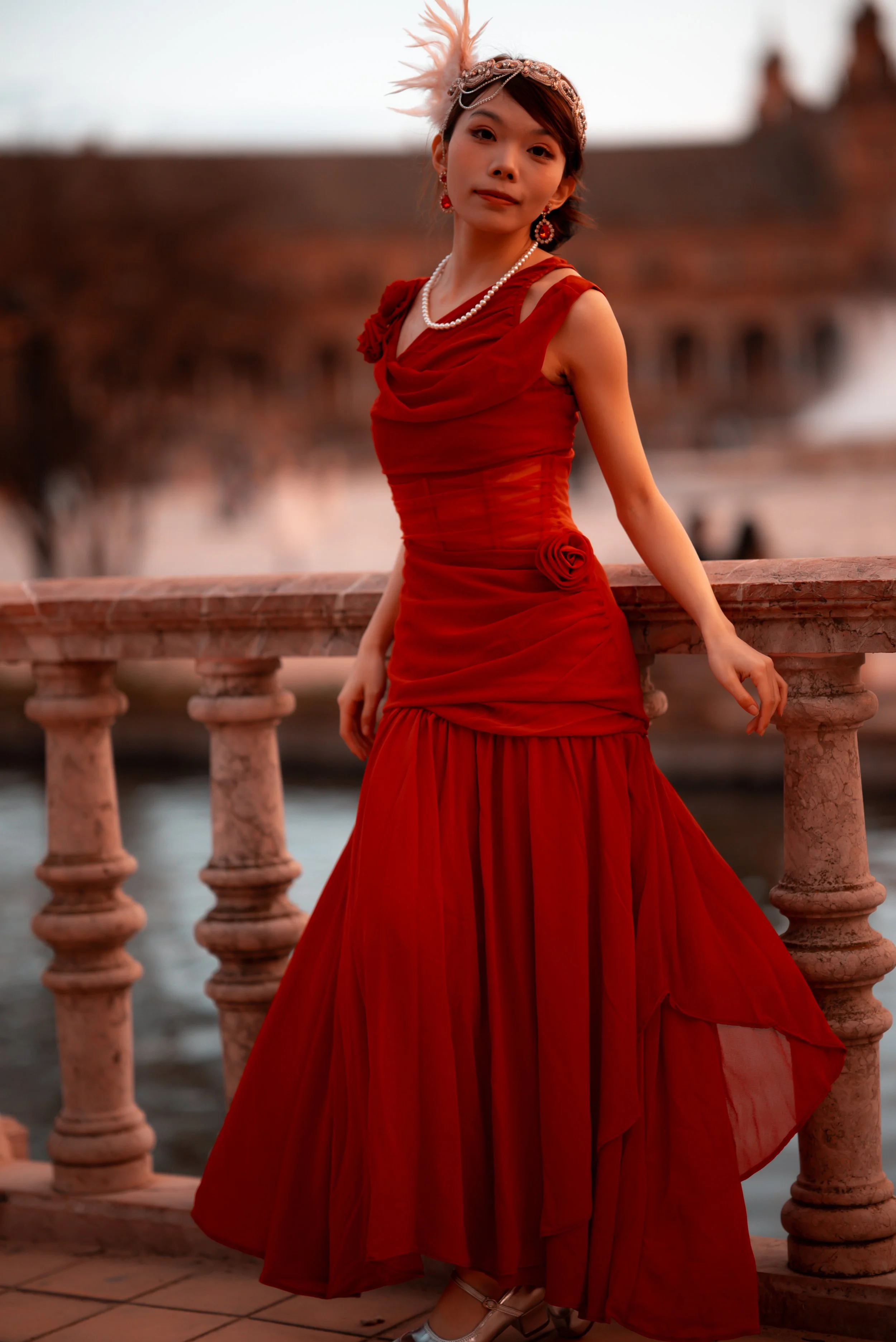 A woman in a red evening gown with decorative flowers and a pleated skirt, wearing pearl jewelry and a feathered headpiece, standing outdoors on a balcony with a river and architecture in the background.
