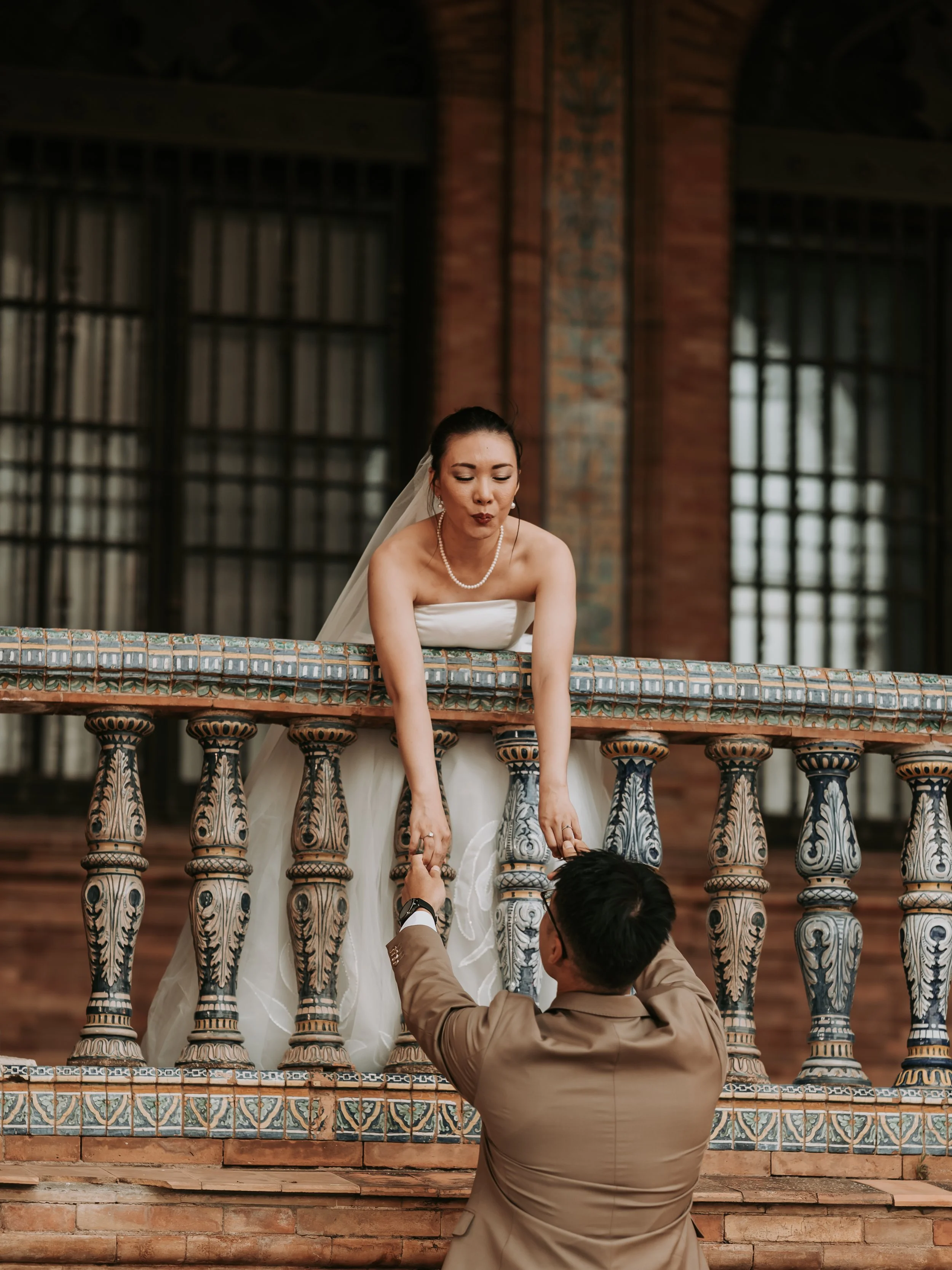 A bride in a white strapless wedding dress and pearl necklace is leaning over a decorative brick and tile railing, holding hands with a man in a beige suit who is kneeling below her. The man is reaching up to her, holding her hands. The background fe