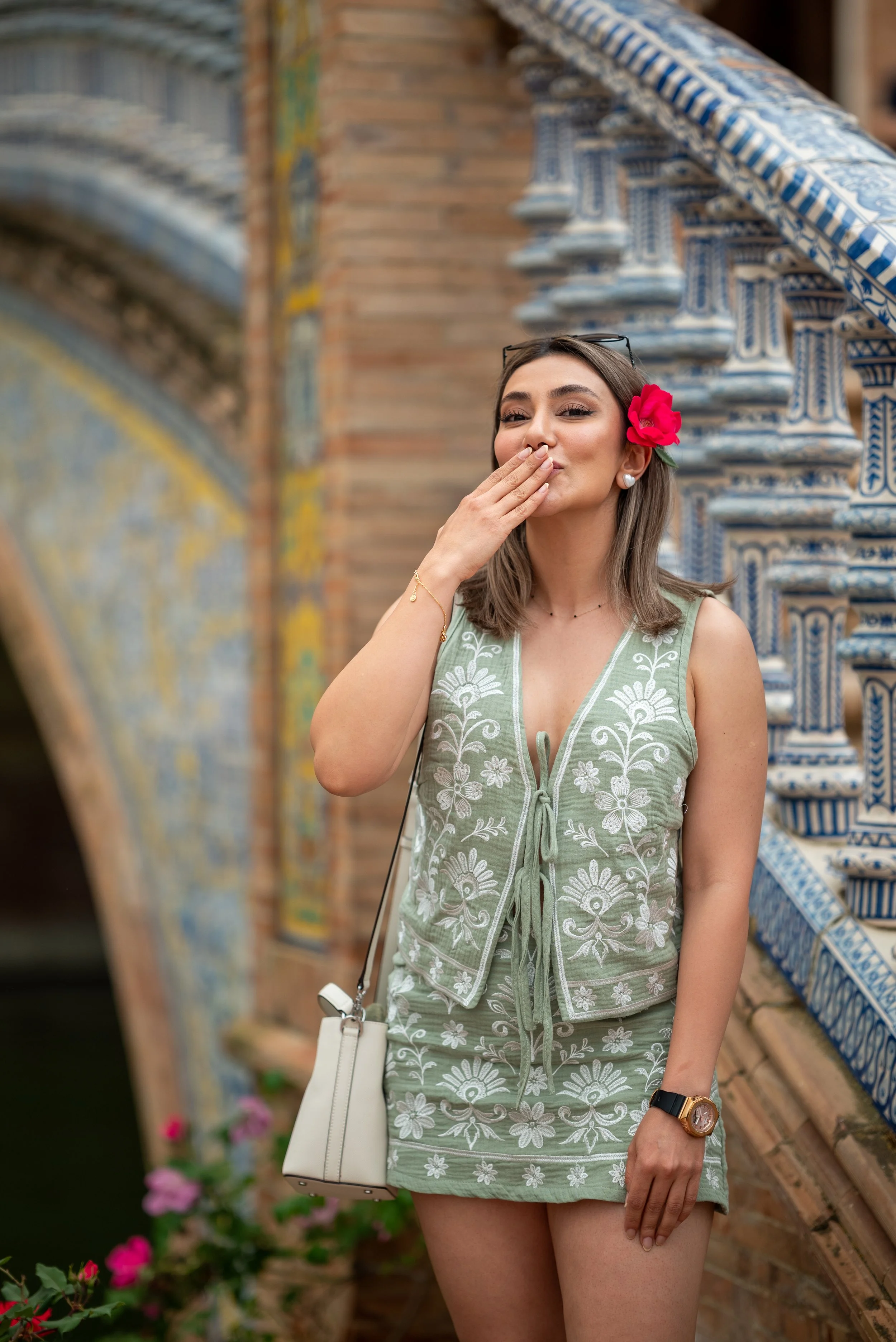 A woman with a pink flower in her hair is standing outdoors near a brick and tile wall. She is wearing a green embroidered dress with floral patterns, a white crossbody purse, sunglasses on her head, a gold bracelet, a black watch, and pearl earrings