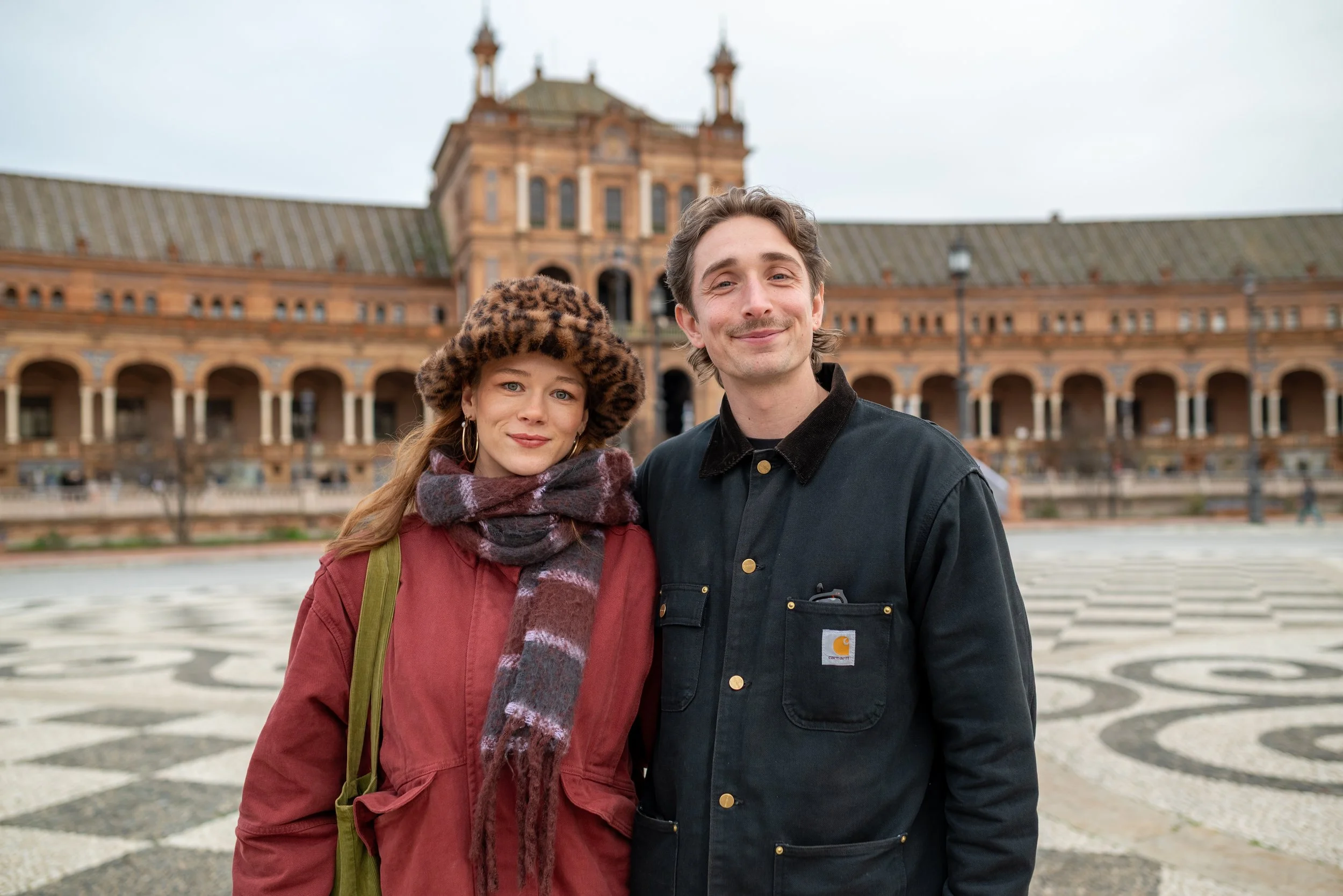 A young woman wearing a leopard-print hat and scarf, and a young man with brown hair and a black Carhartt jacket, standing in front of a historic brick building with arches and towers.
