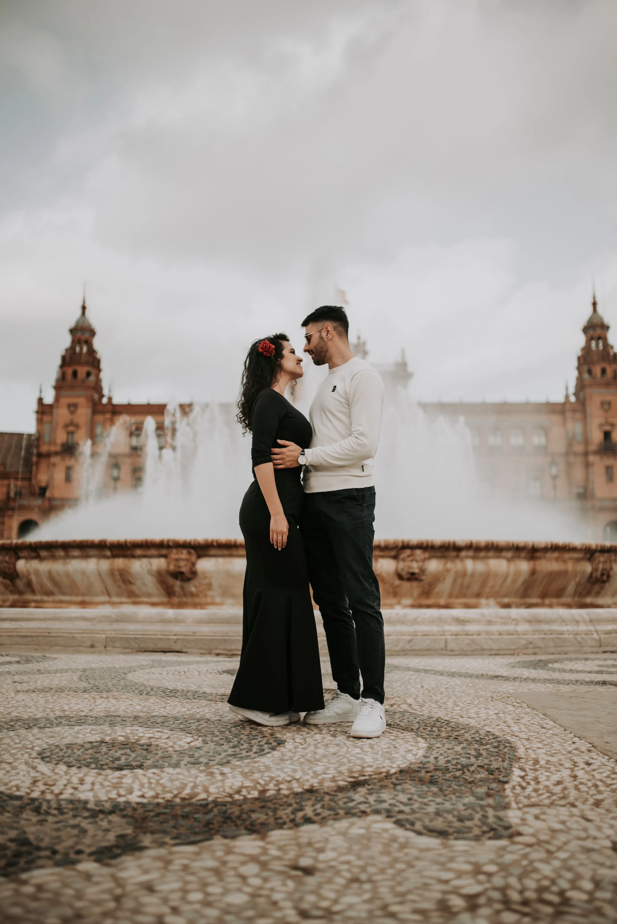 A couple stands close together, faces nearly touching, in front of a historic building with fountains. The woman has long curly hair with a red flower, and the man has short dark hair, sunglasses, and white sneakers.