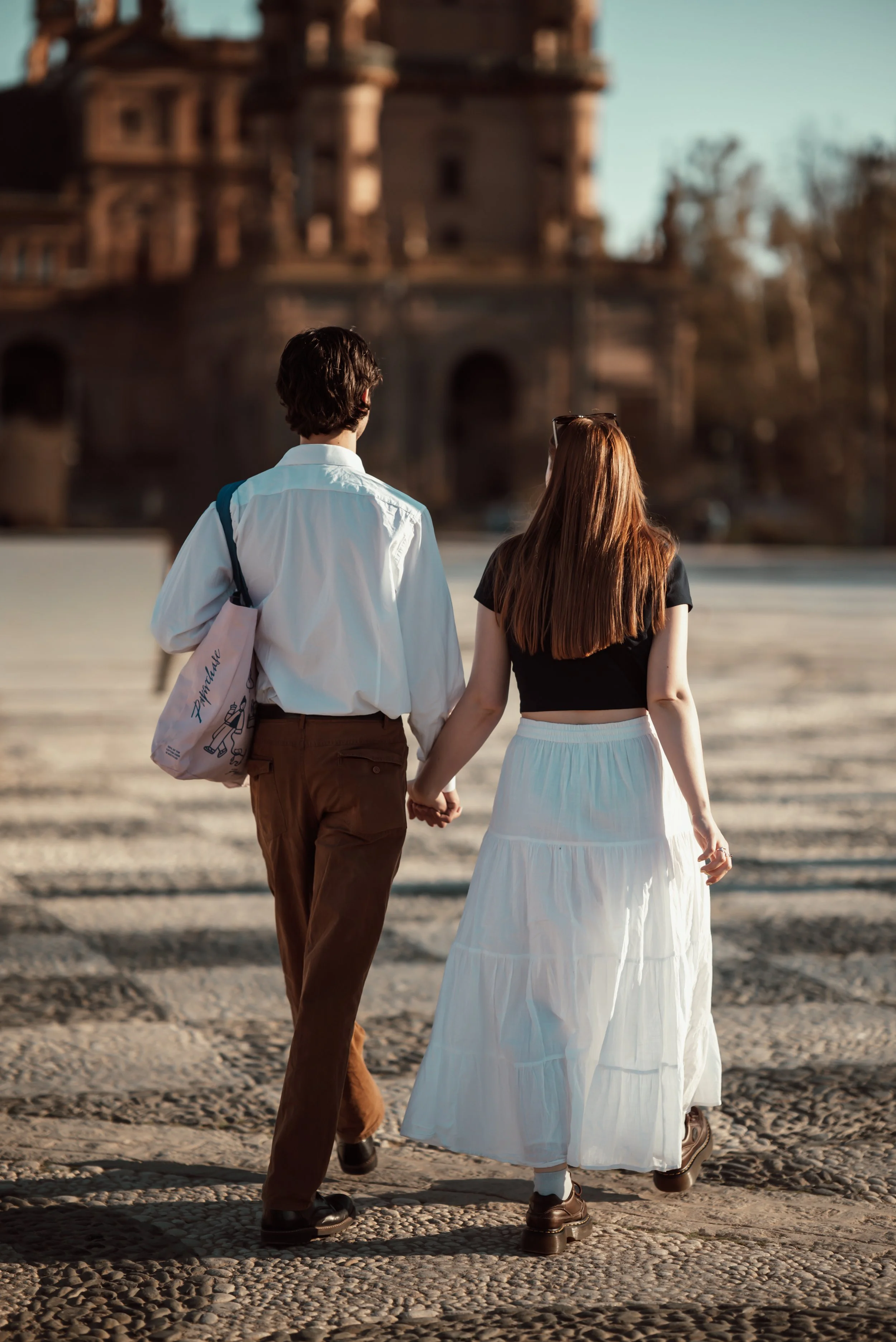 A young man and woman holding hands and walking away from the camera on a cobblestone street. The man wears brown pants, a white shirt, and carries a tote bag. The woman wears a white skirt and a black top. They are walking towards a blurred historic