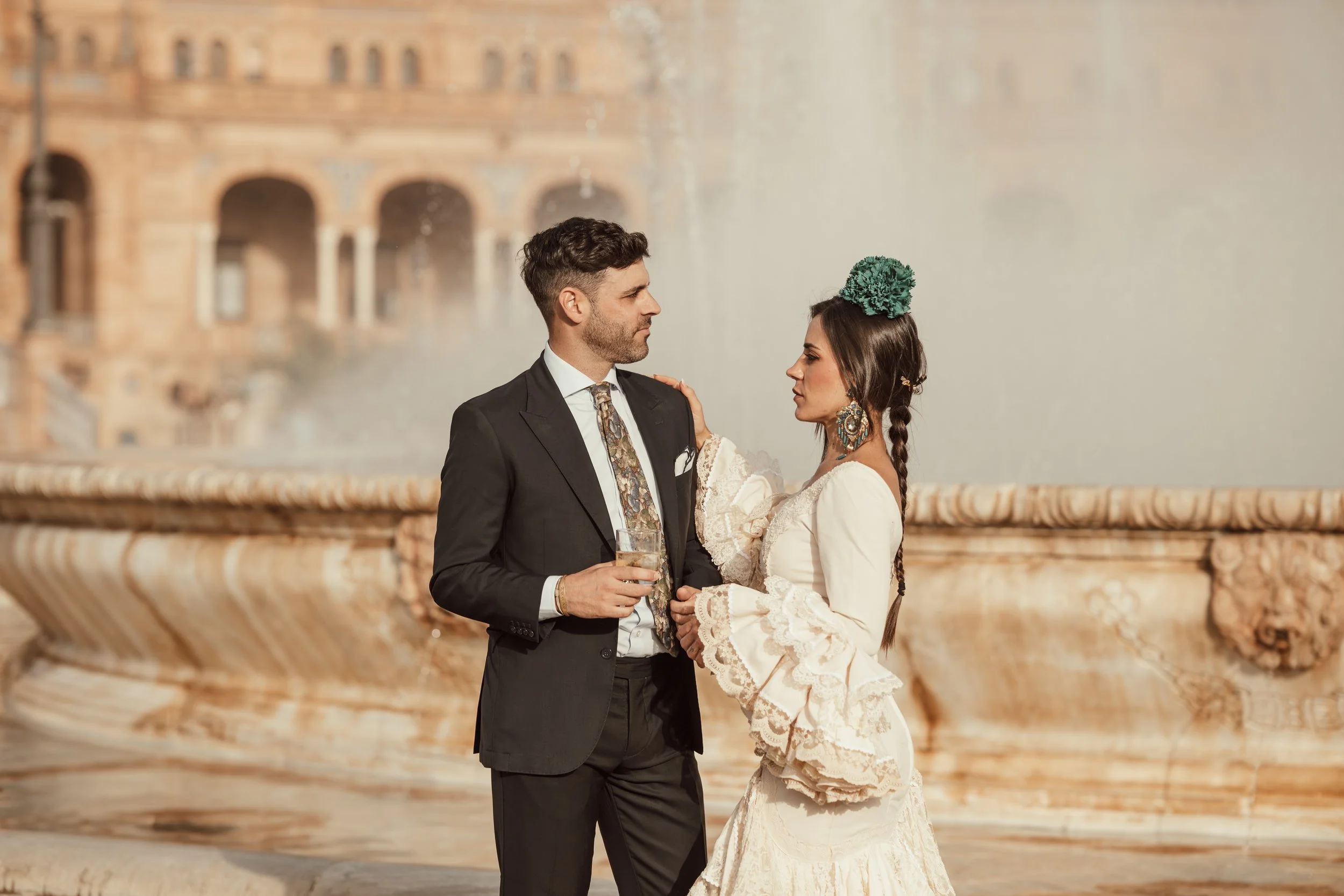 A man in a black suit and a woman in a white lace dress are standing close together in front of a fountain with water spraying behind them. The man is holding a drink, and both are looking at each other. The woman is wearing large earrings and a teal