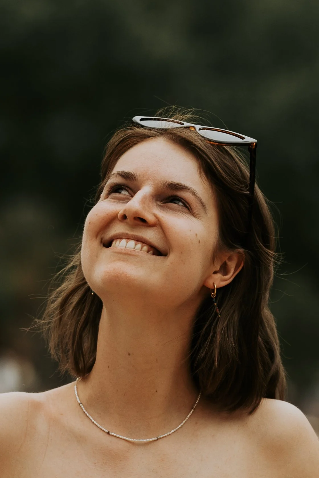 Smiling woman with brown hair and sunglasses on her head, wearing a necklace and earrings, looking upward.