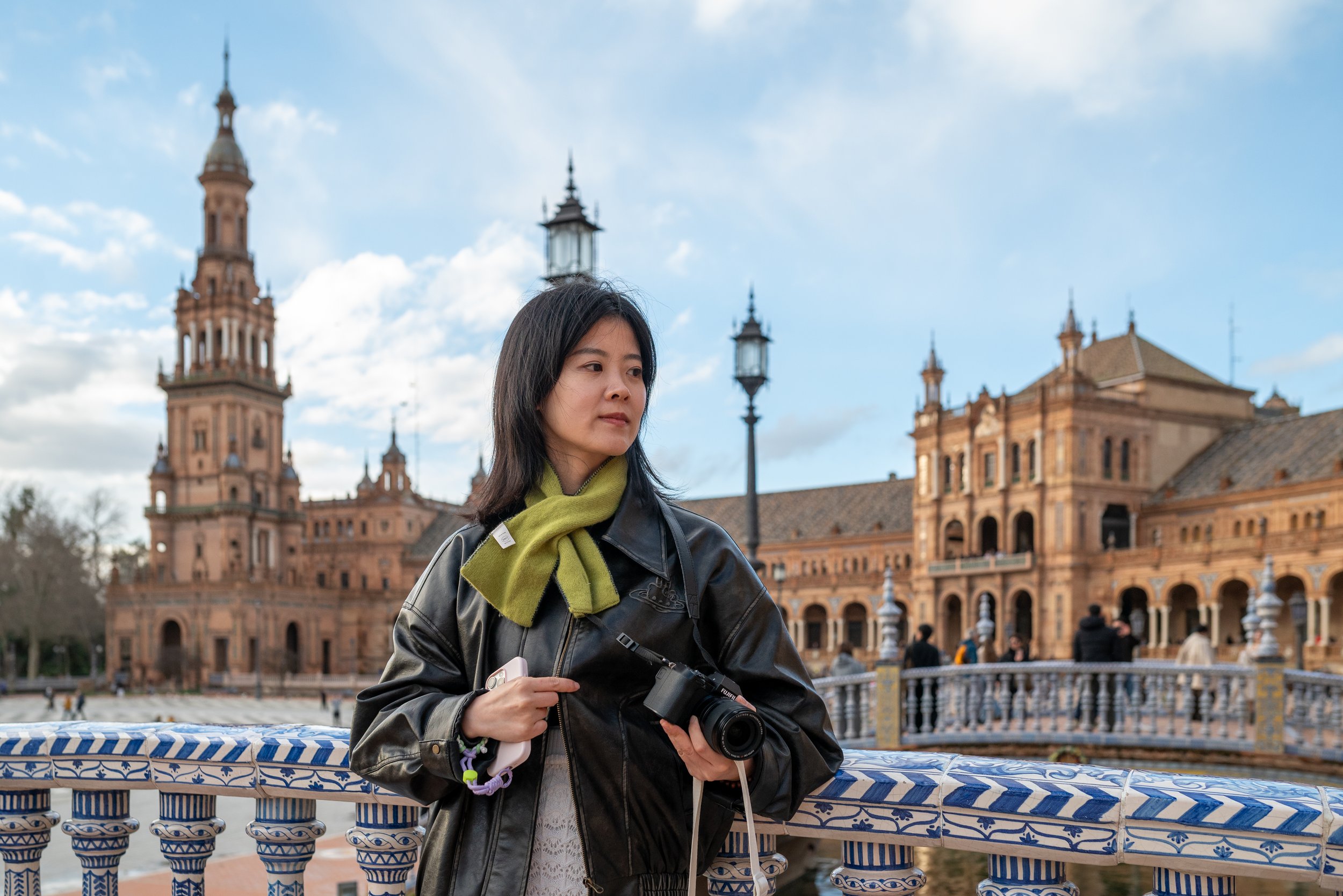 A woman standing on a bridge holding a camera, with historic buildings and a cloudy sky in the background.