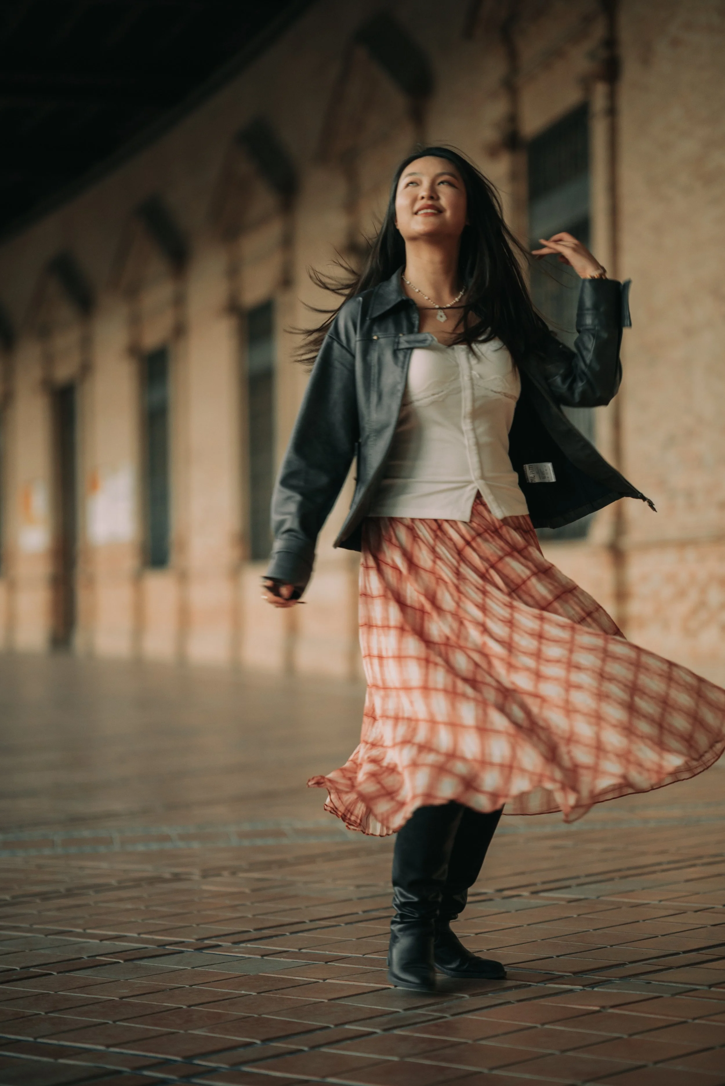 A woman with long dark hair smiling and twirling in a flowing orange and white patterned skirt, black boots, a white top, a black leather jacket, and jewelry, outdoors on a brick sidewalk at dusk.
