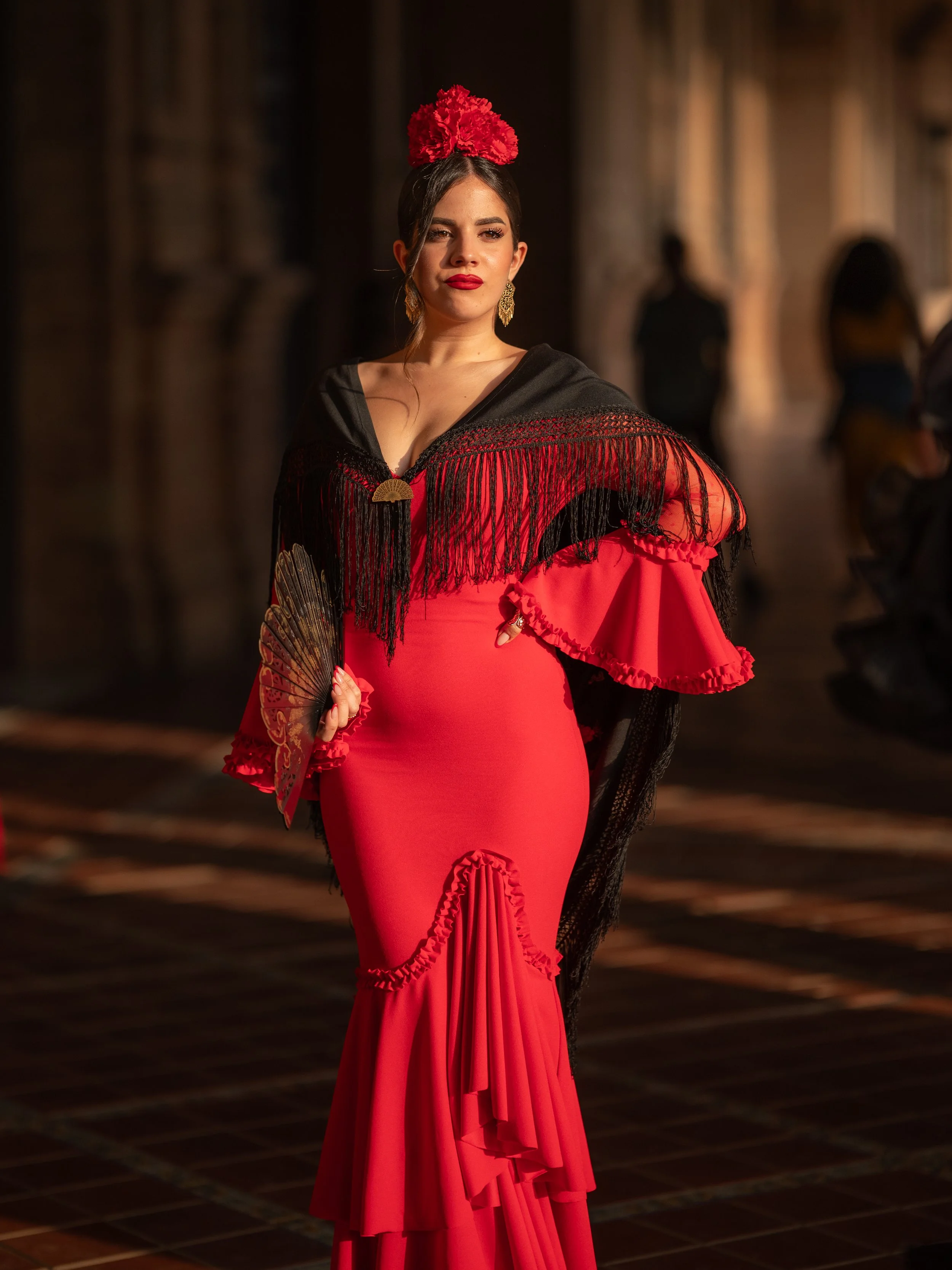 A woman dressed as a traditional Spanish flamenco dancer, wearing a red dress with ruffles, a black shawl with fringe, and a red flower headpiece, holding a fan, on a dimly lit street at night.