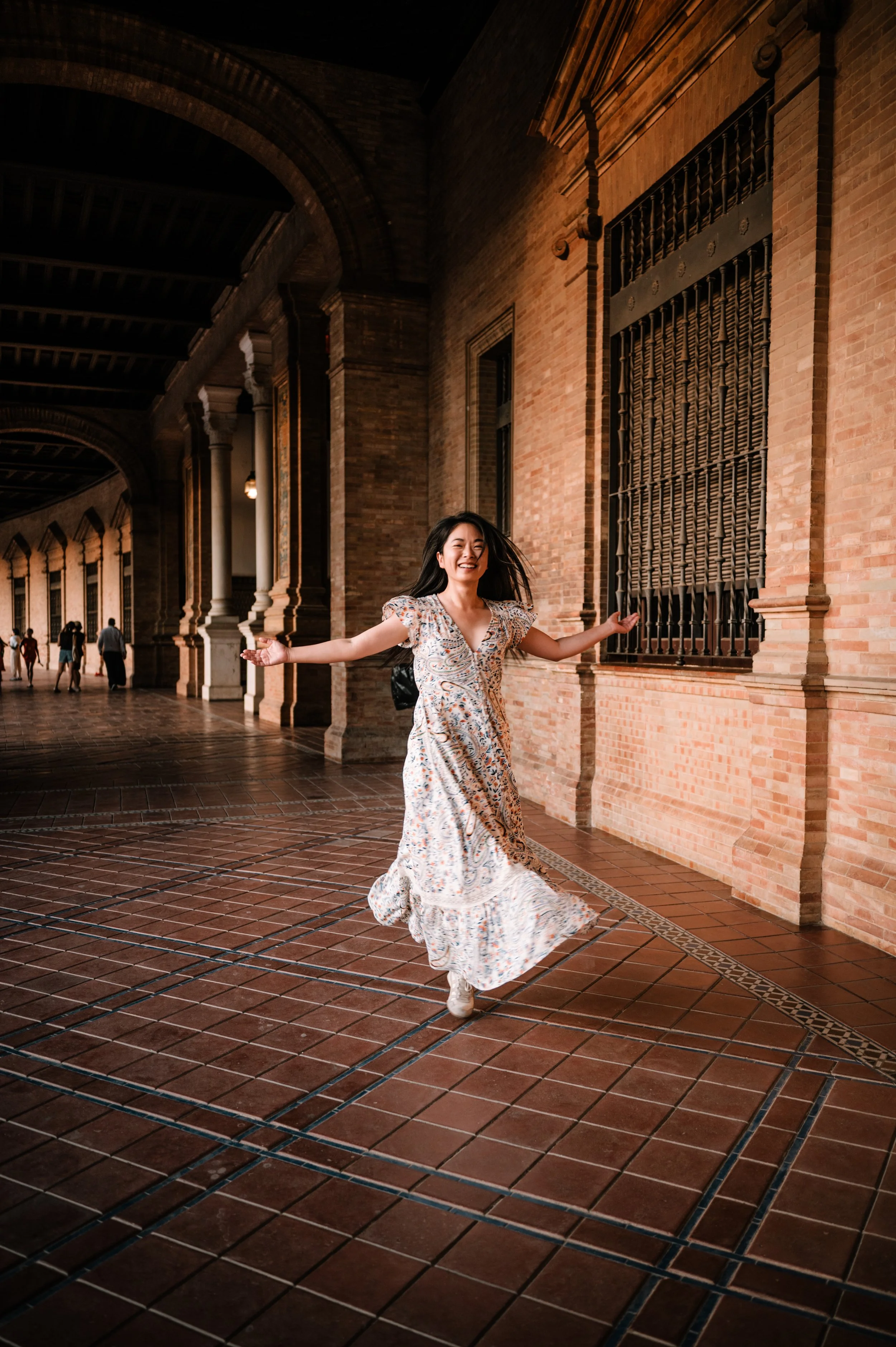 A woman in a floral dress smiling and spinning with arms outstretched in a historic brick arcade or courtyard.