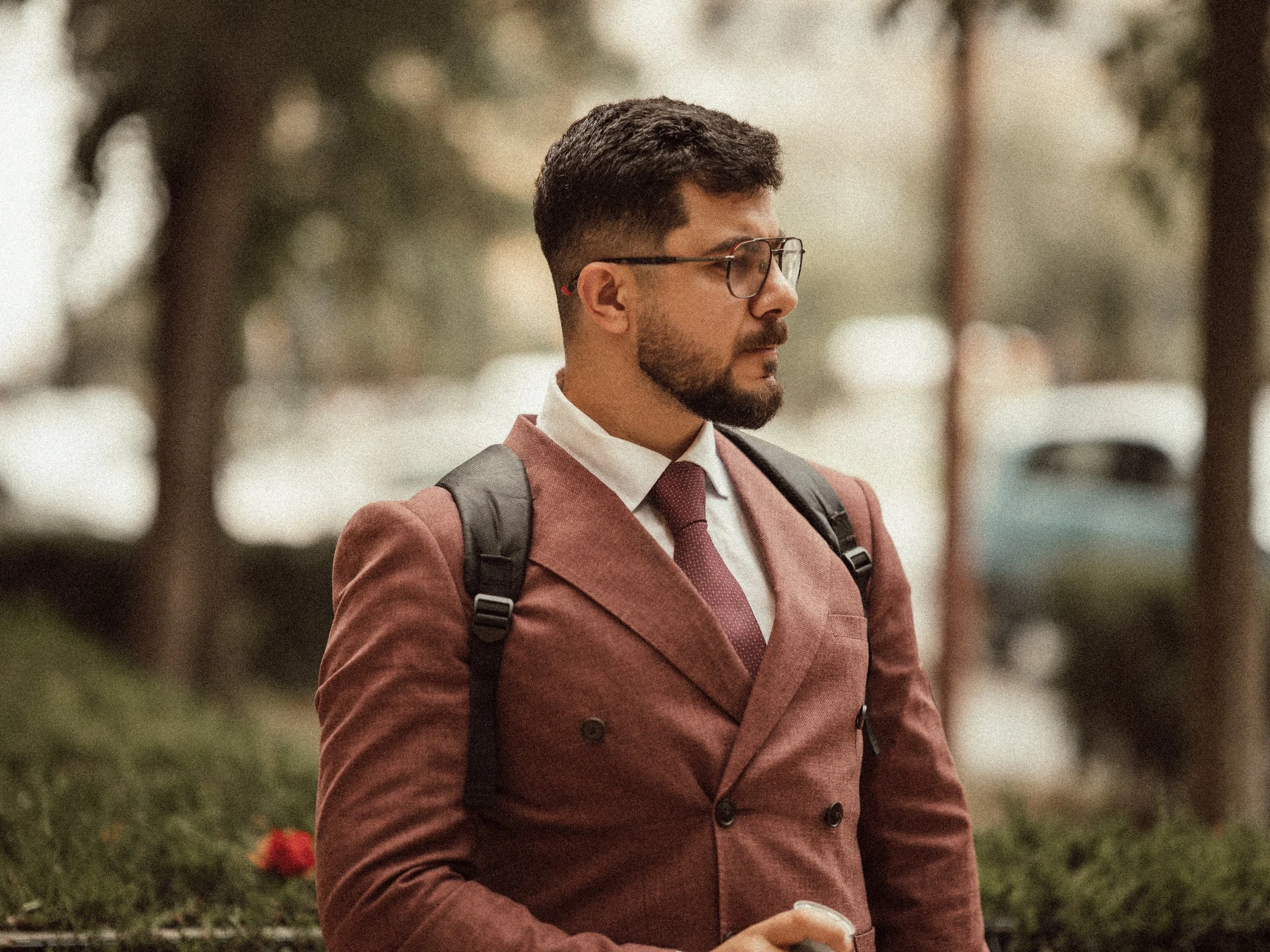 Man with short dark hair, glasses, and beard wearing a pink suit and tie, carrying a backpack, outdoors with blurred trees and flowers in the background.