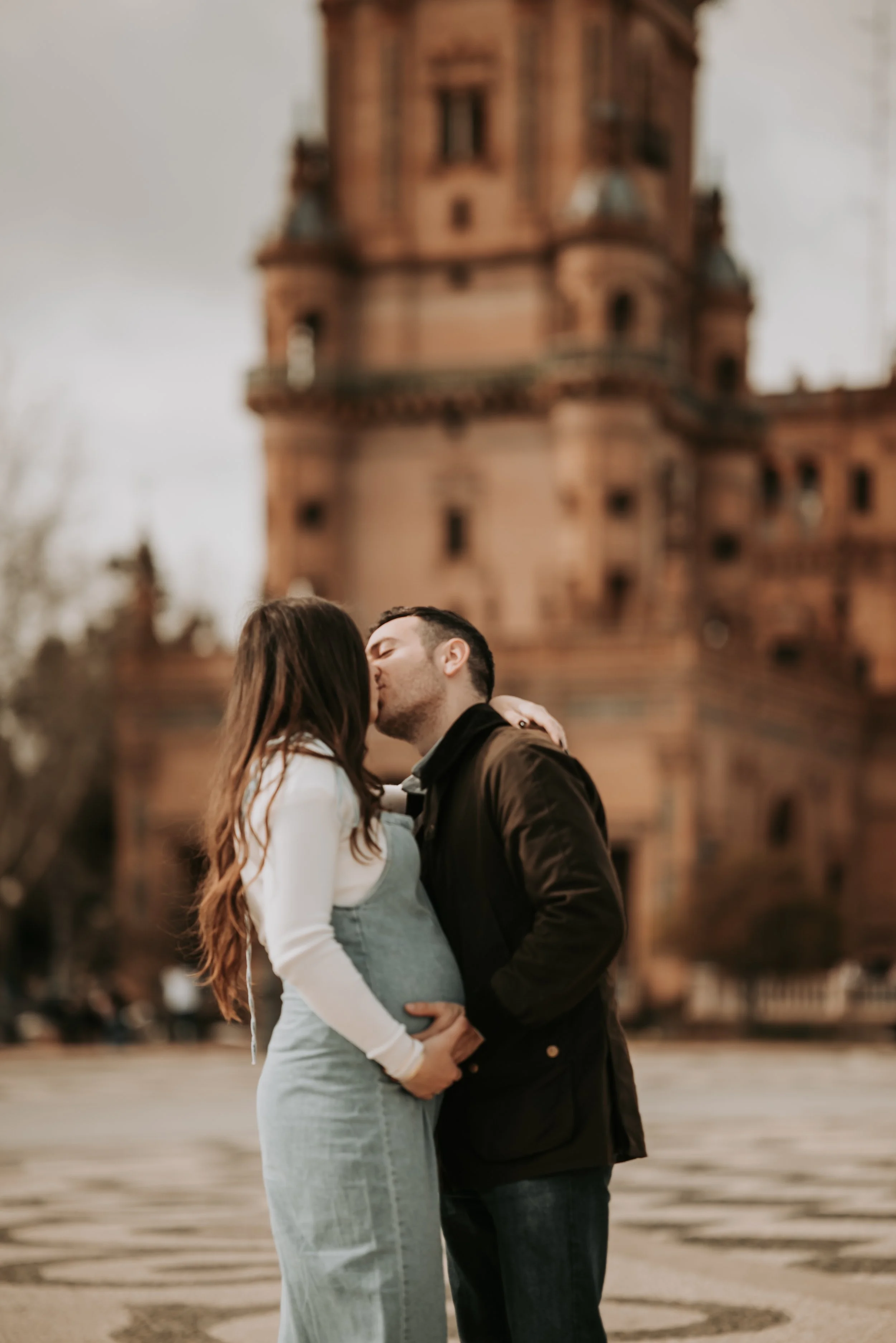 A couple sharing a kiss in front of a historic castle, with the woman visibly pregnant, standing close together with the man's hand on her belly, during evening or dusk.