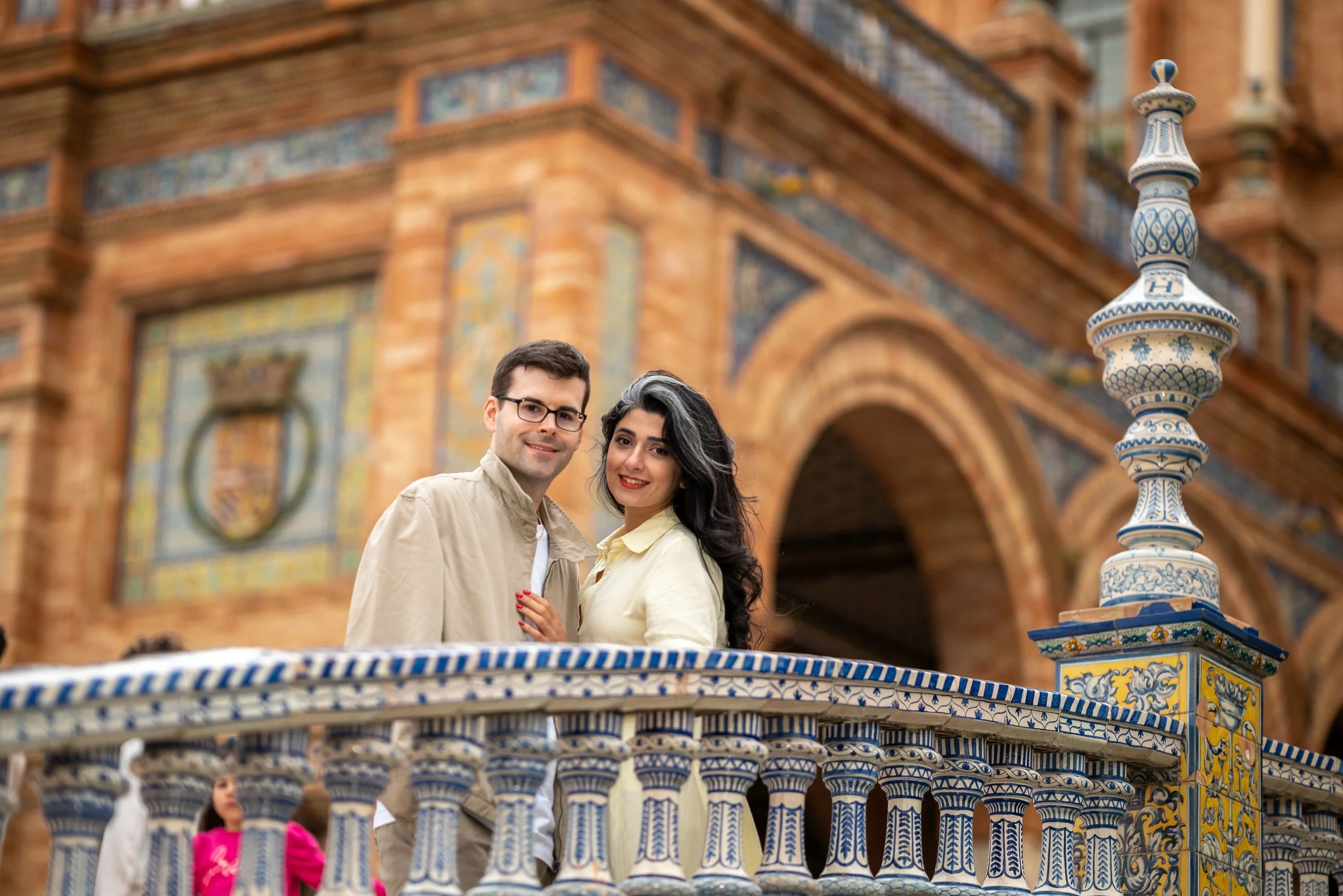 A couple stands on a decorative carousel with ornate blue, yellow, and white tiling in front of a historic brick building with decorative tiles and an archway.
