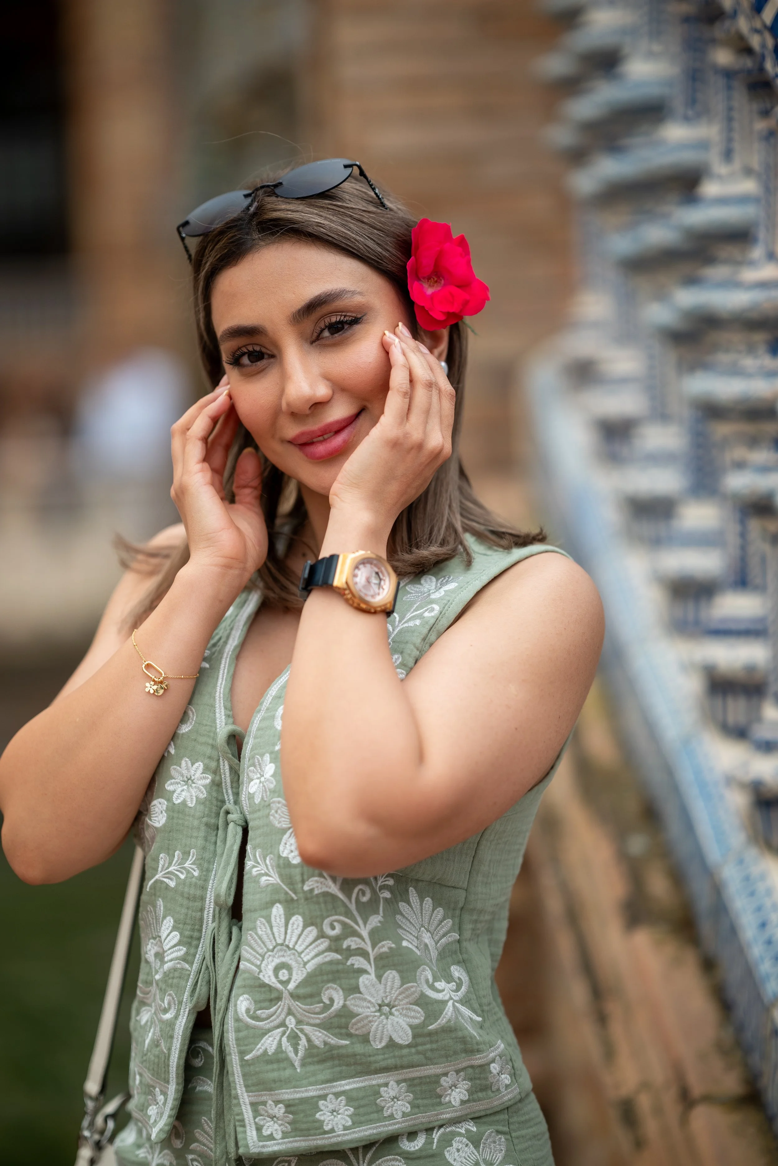 A woman with brown hair wearing sunglasses on her head, a pink flower placed near her ear, a gold watch, and a gold bracelet, posing in front of a row of wine bottles.