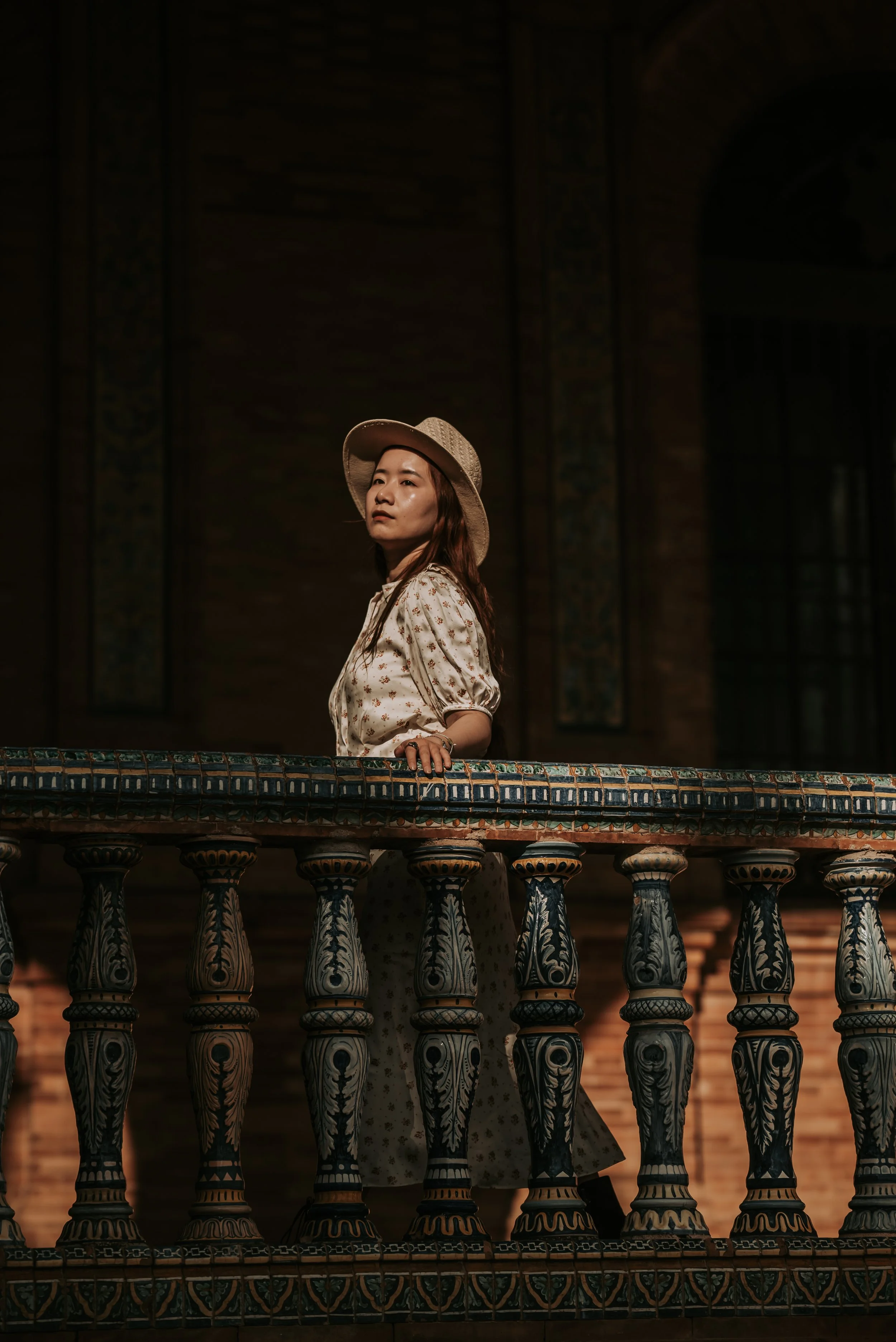 A woman with long hair wearing a hat and patterned dress standing on a balcony with ornate railing, looking contemplative, at dusk or night.
