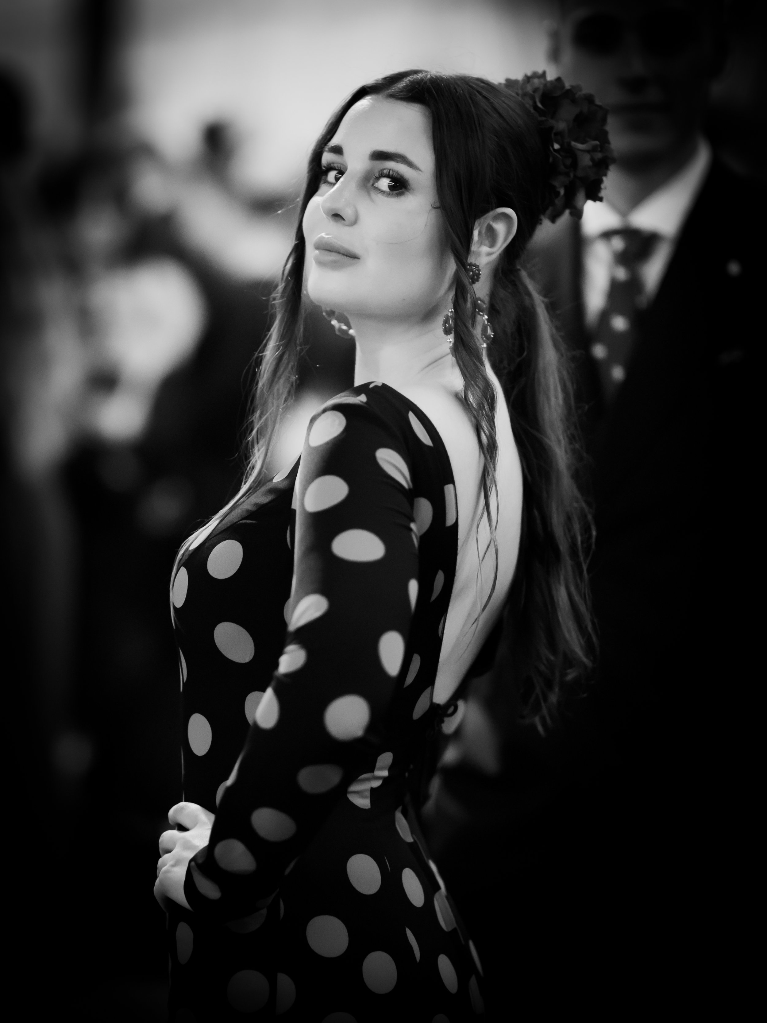 A woman dressed in a polka dot dress with a low back, wearing earrings and with her hair styled with flowers, looking over her shoulder in a black and white photo at a social event.