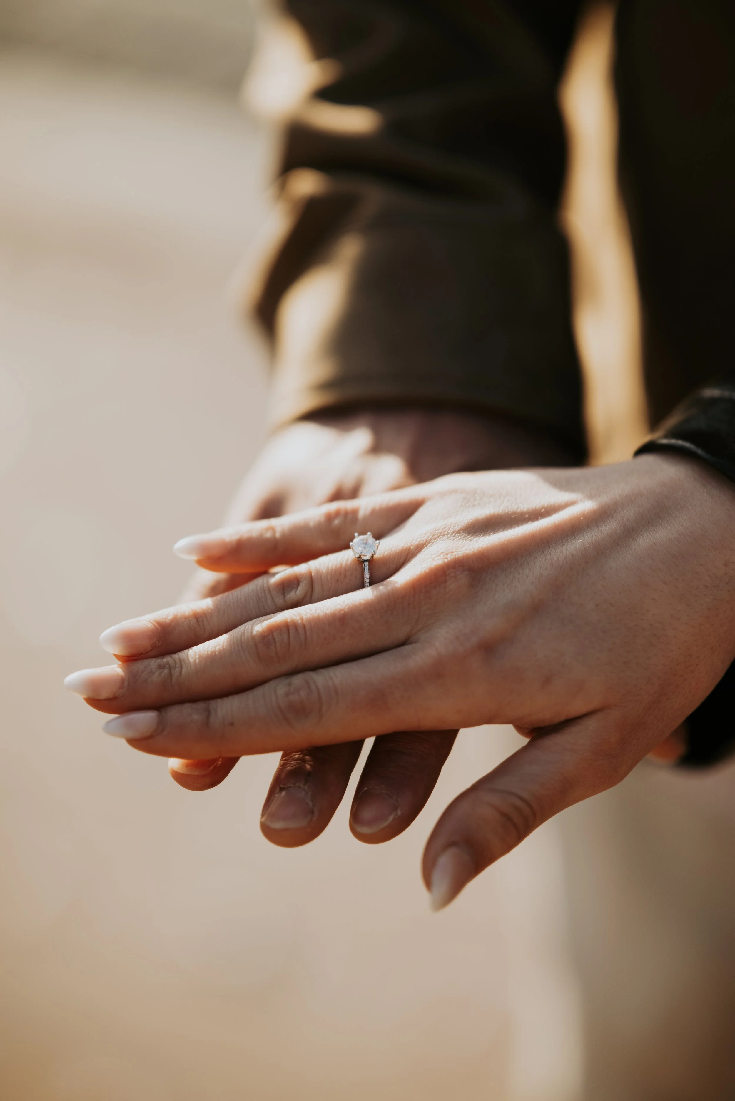 Close-up of a woman showing her left hand with an engagement ring on her ring finger, with a man’s hand holding her hand from below.