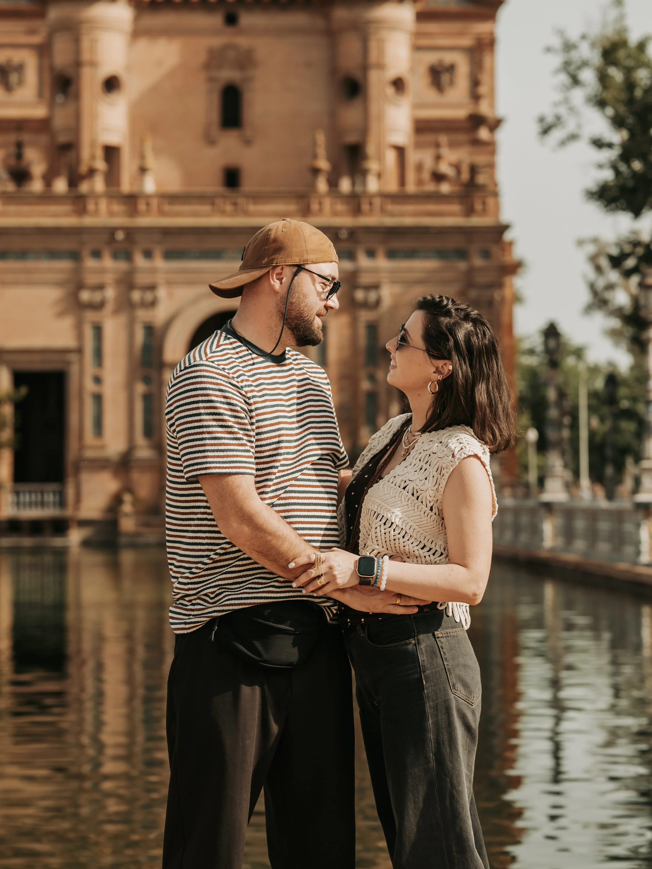 A couple standing close together near a body of water with a historic building in the background. They are smiling and looking at each other, holding hands.
