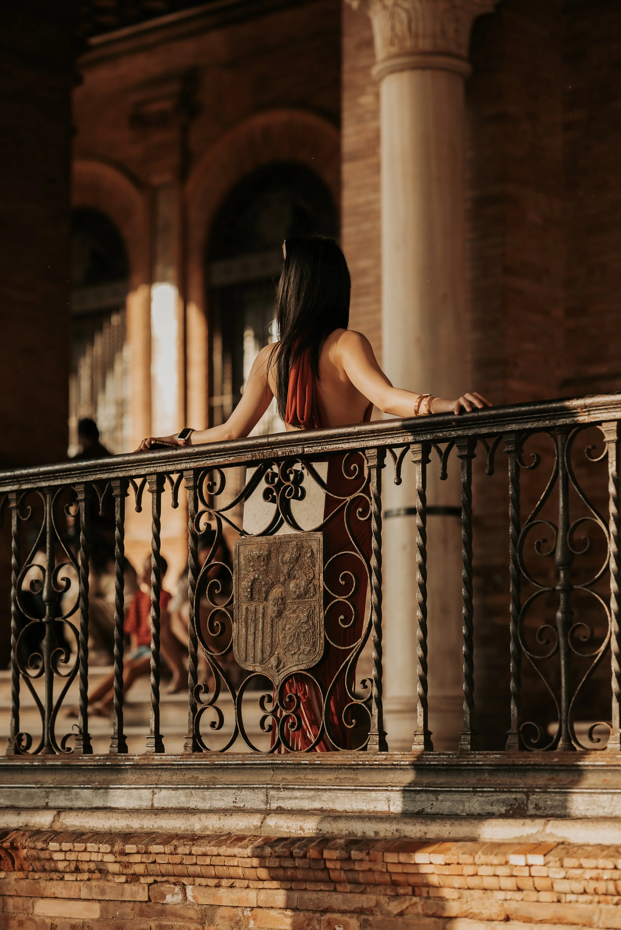 A woman with long black hair wearing a red dress standing on a balcony with ornate black iron railing inside a historic brick building with large columns and arched windows.