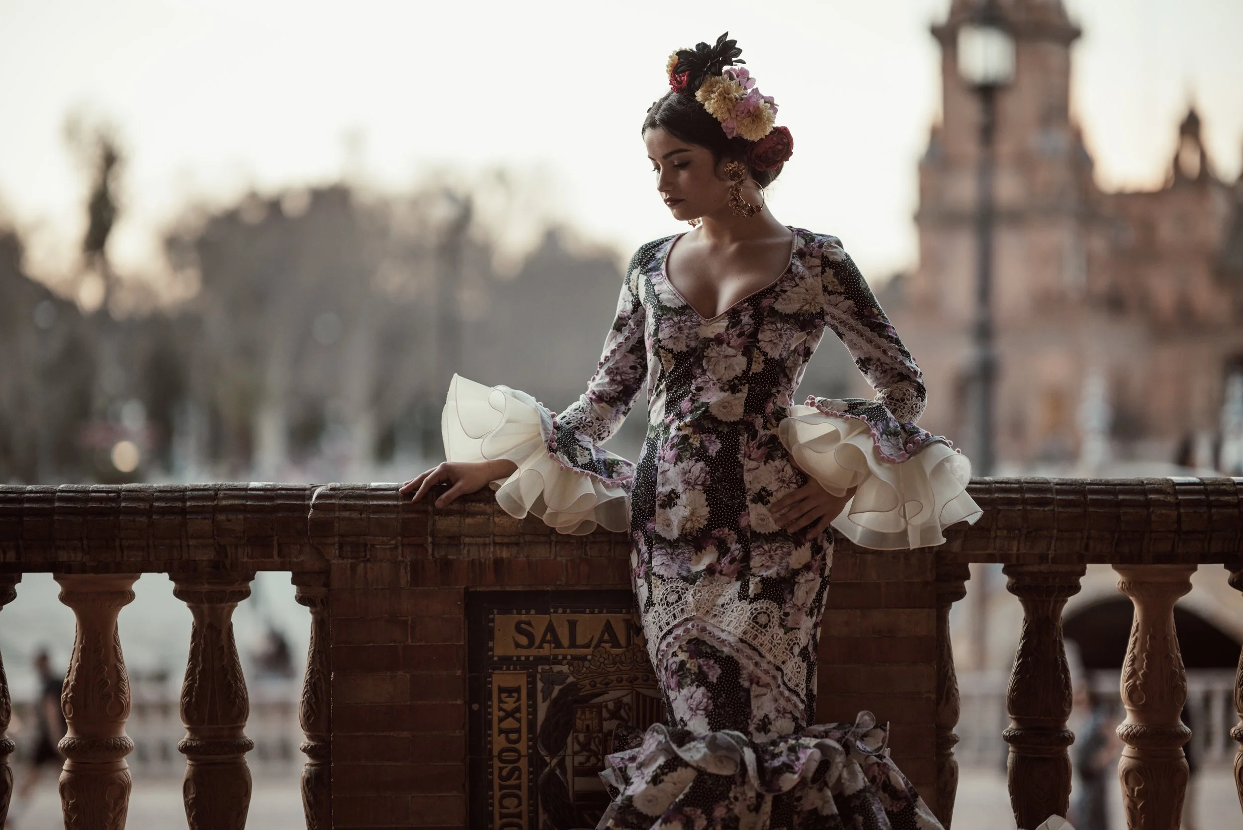 A woman dressed in traditional Mexican attire with a floral shawl and large earrings, standing on a balcony with ornate balusters. She has a floral headpiece and appears to be in Mexico City, possibly in a historic setting.