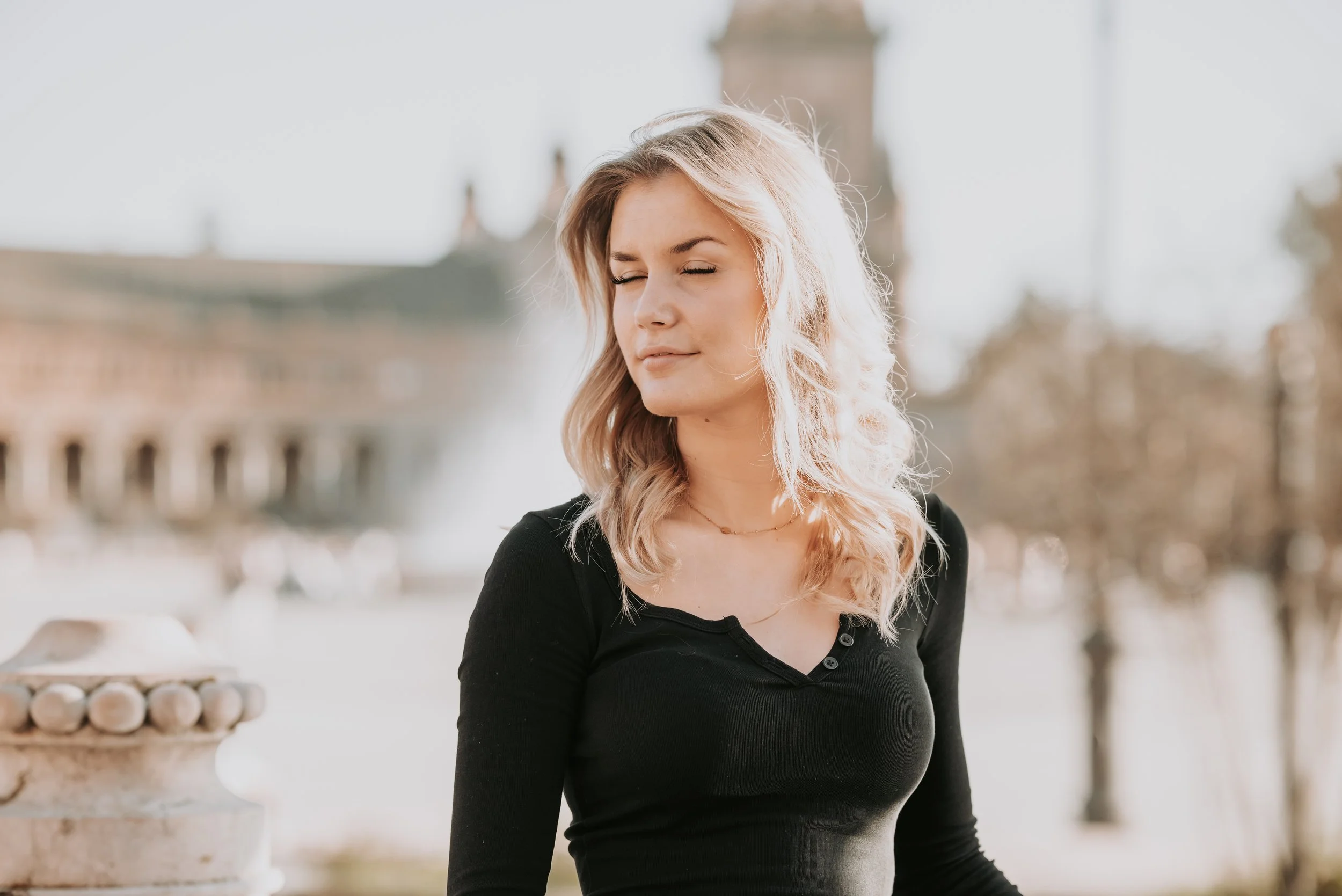 A young woman with blonde wavy hair and closed eyes stands outdoors in front of a blurred background with a stone balustrade and fountain.