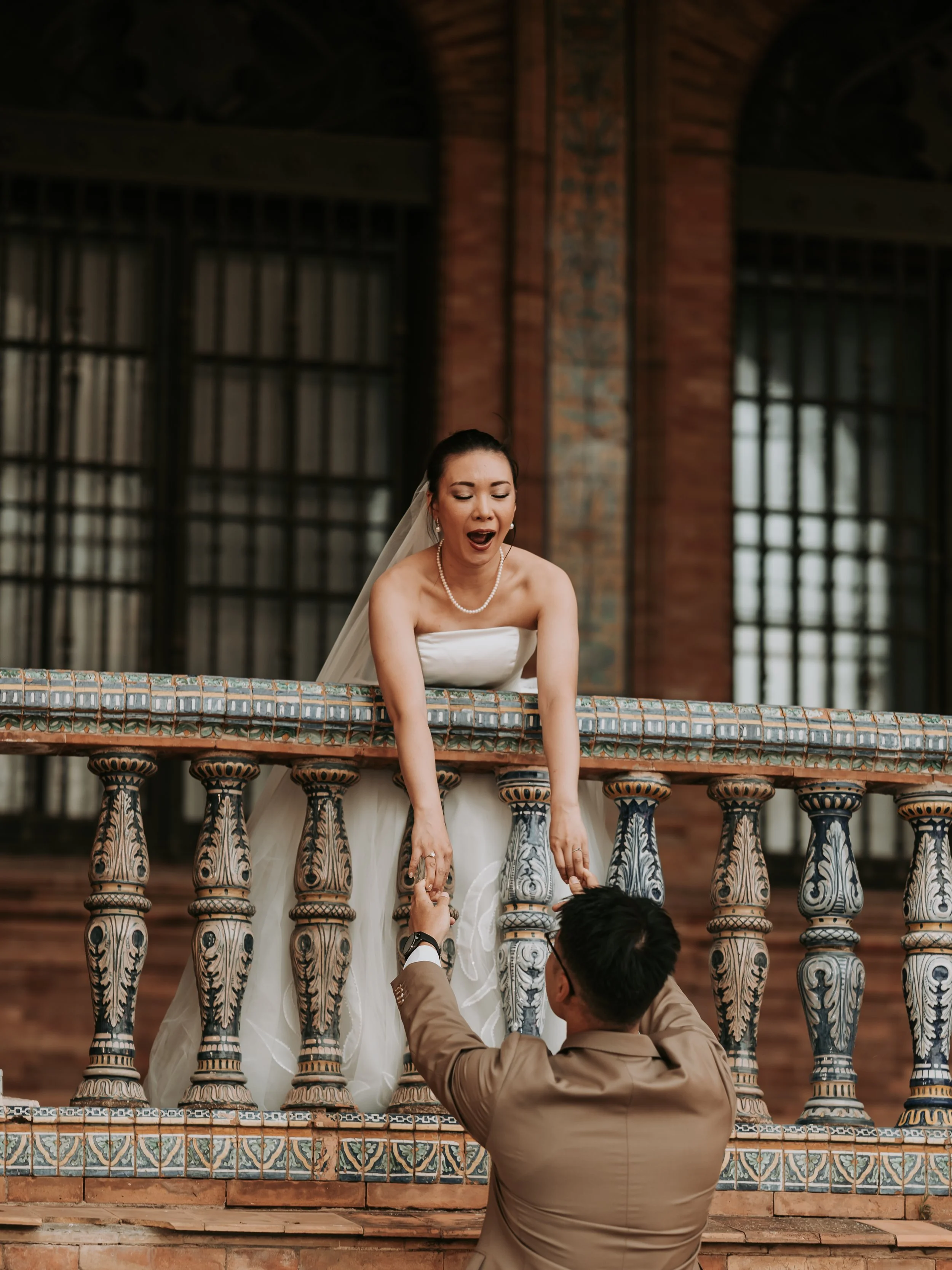 A bride in a white wedding dress and pearl necklace is leaning over a decorative balcony, smiling with her eyes closed. She is holding the hands of a man dressed in a beige suit, who is looking up at her. The setting features large windows with iron 