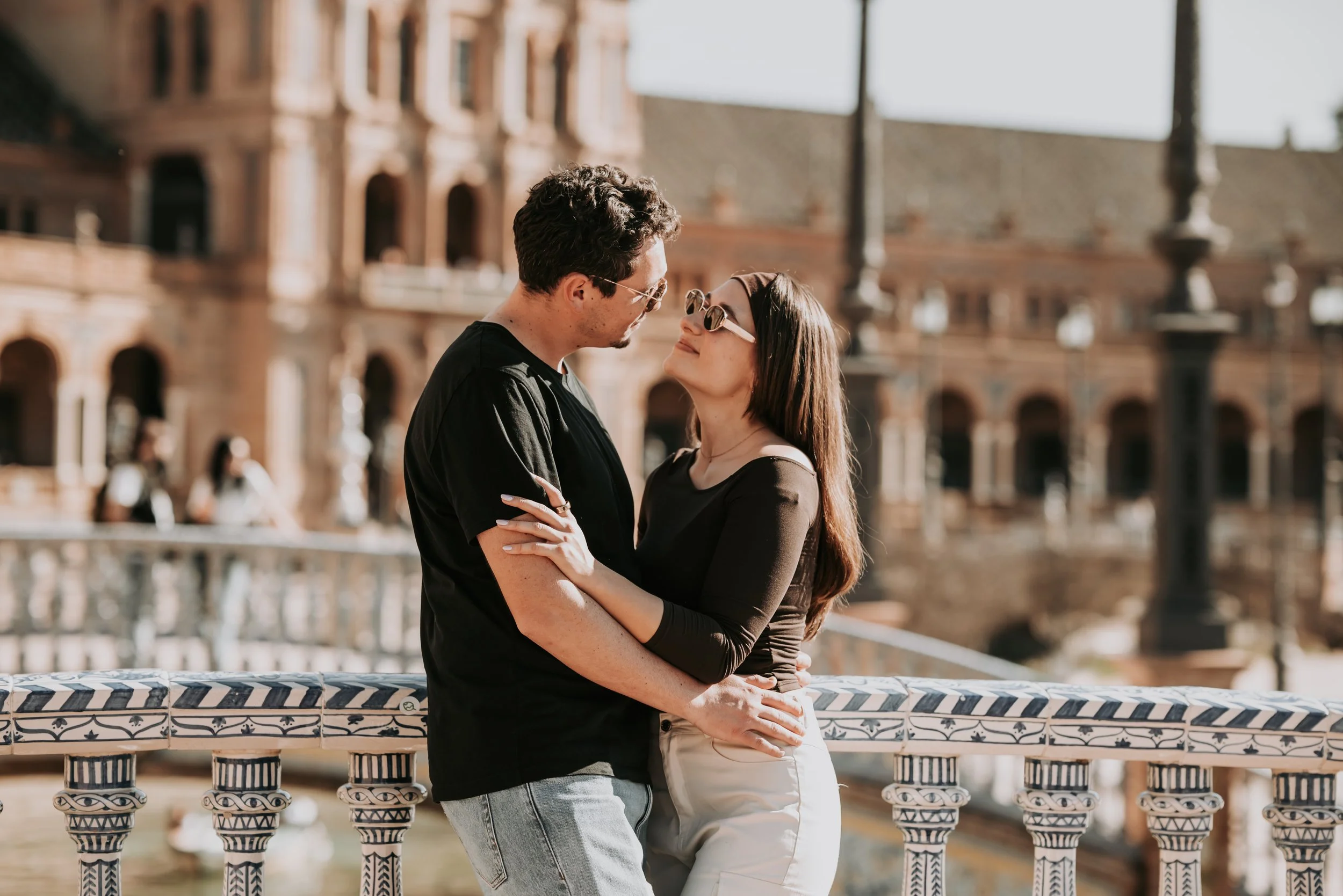 A couple standing closely together on a bridge in front of historic buildings, with the man wearing glasses and a black T-shirt and the woman with long brown hair and round sunglasses, smiling at each other.