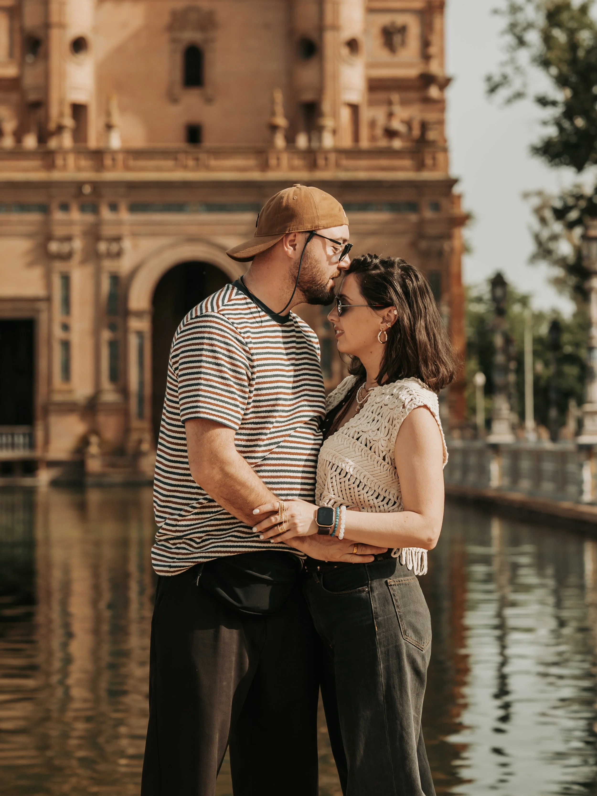 A couple standing close together by a body of water in front of a historic brick building, with trees on the side, sharing an intimate moment.