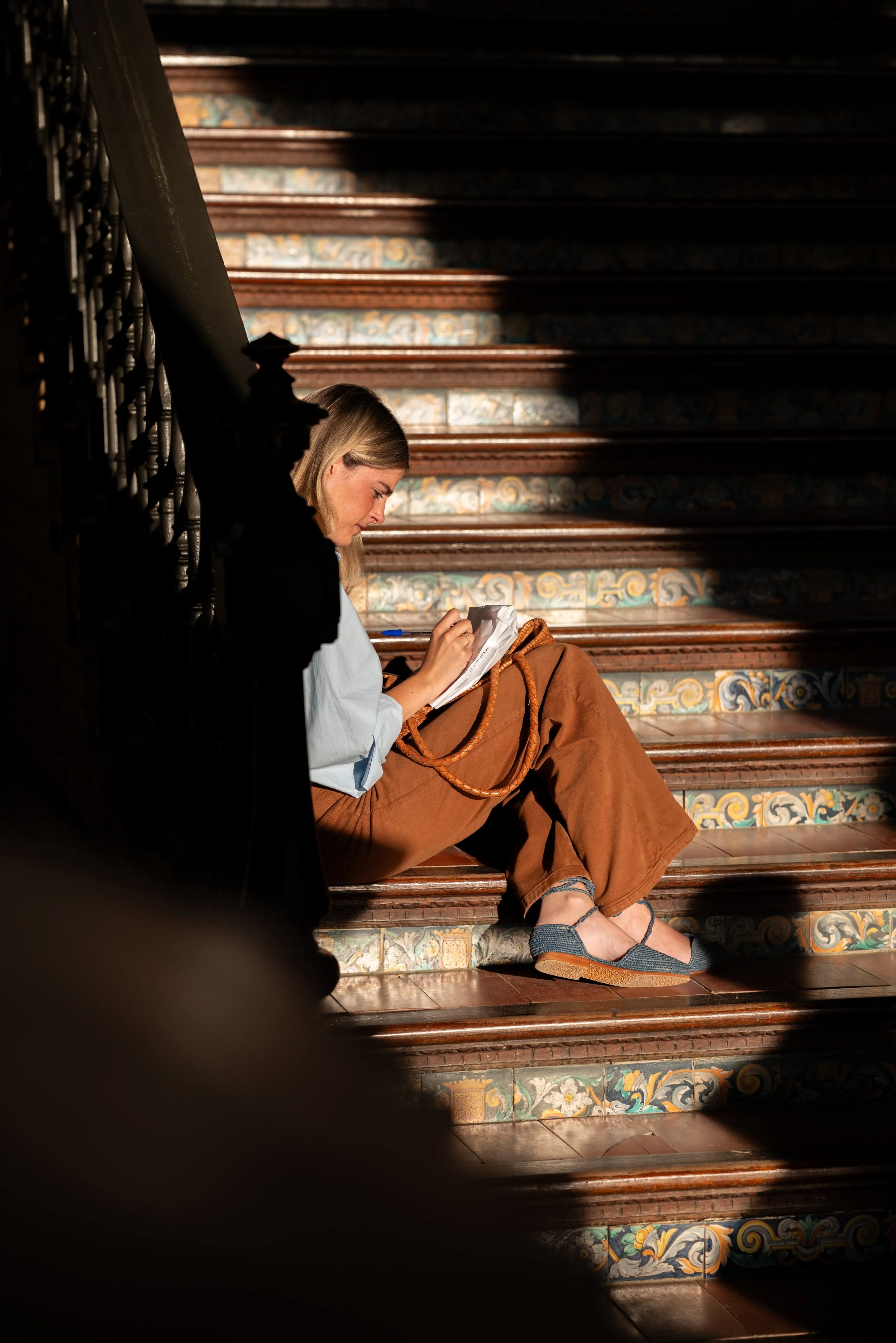 A woman with blonde hair sitting on a decorated staircase, reading or writing in a notebook. She is dressed in a white shirt and brown pants, with denim shoes and a brown bag at her side. The staircase has colorful tiled risers and dark wooden steps.
