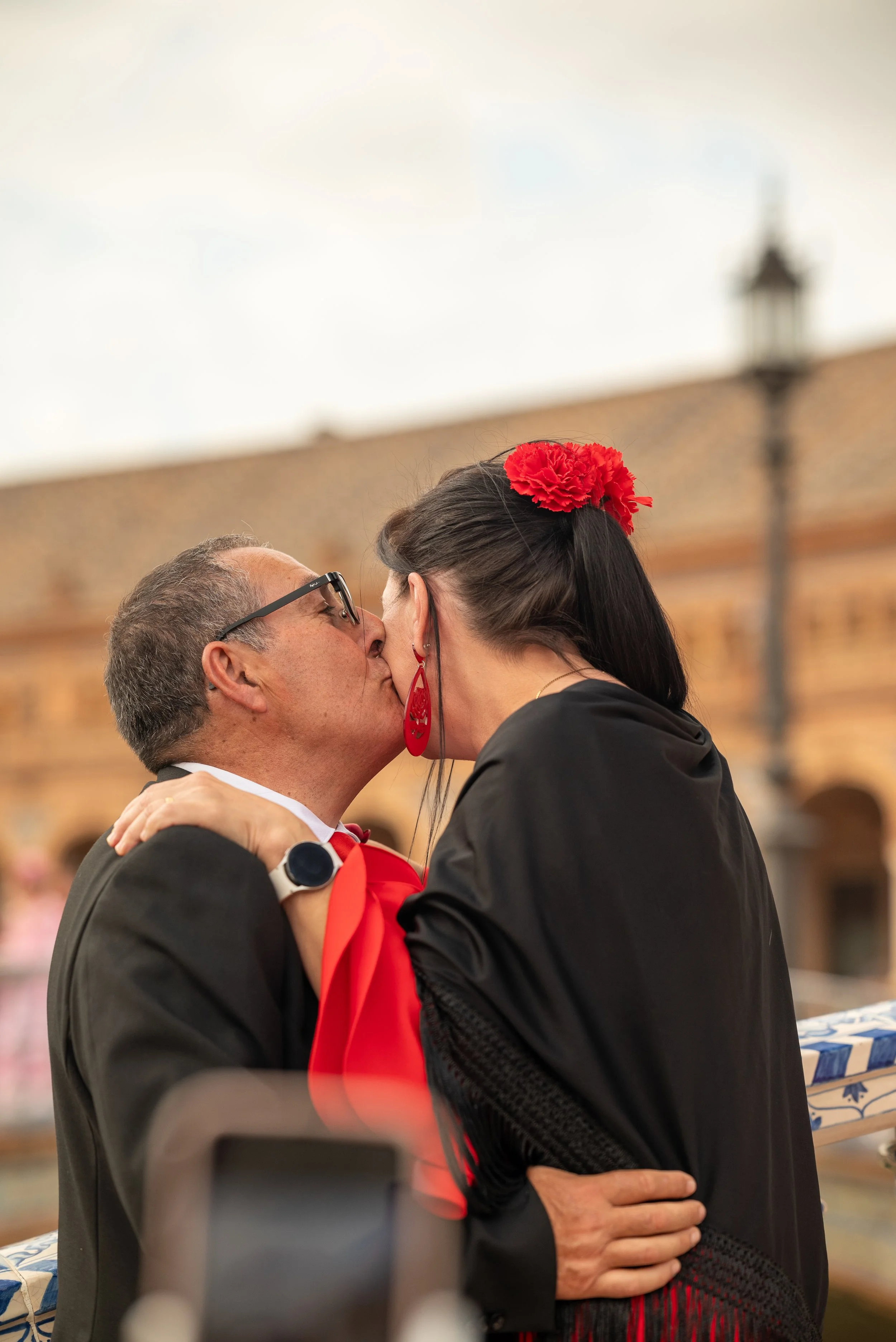 A man and woman share a kiss at a celebration or ceremony. The woman has black hair with a red flower accessory and red jewelry. The man wears glasses and a watch. They are hugging, and the background shows a brick wall and outdoor lamp post.