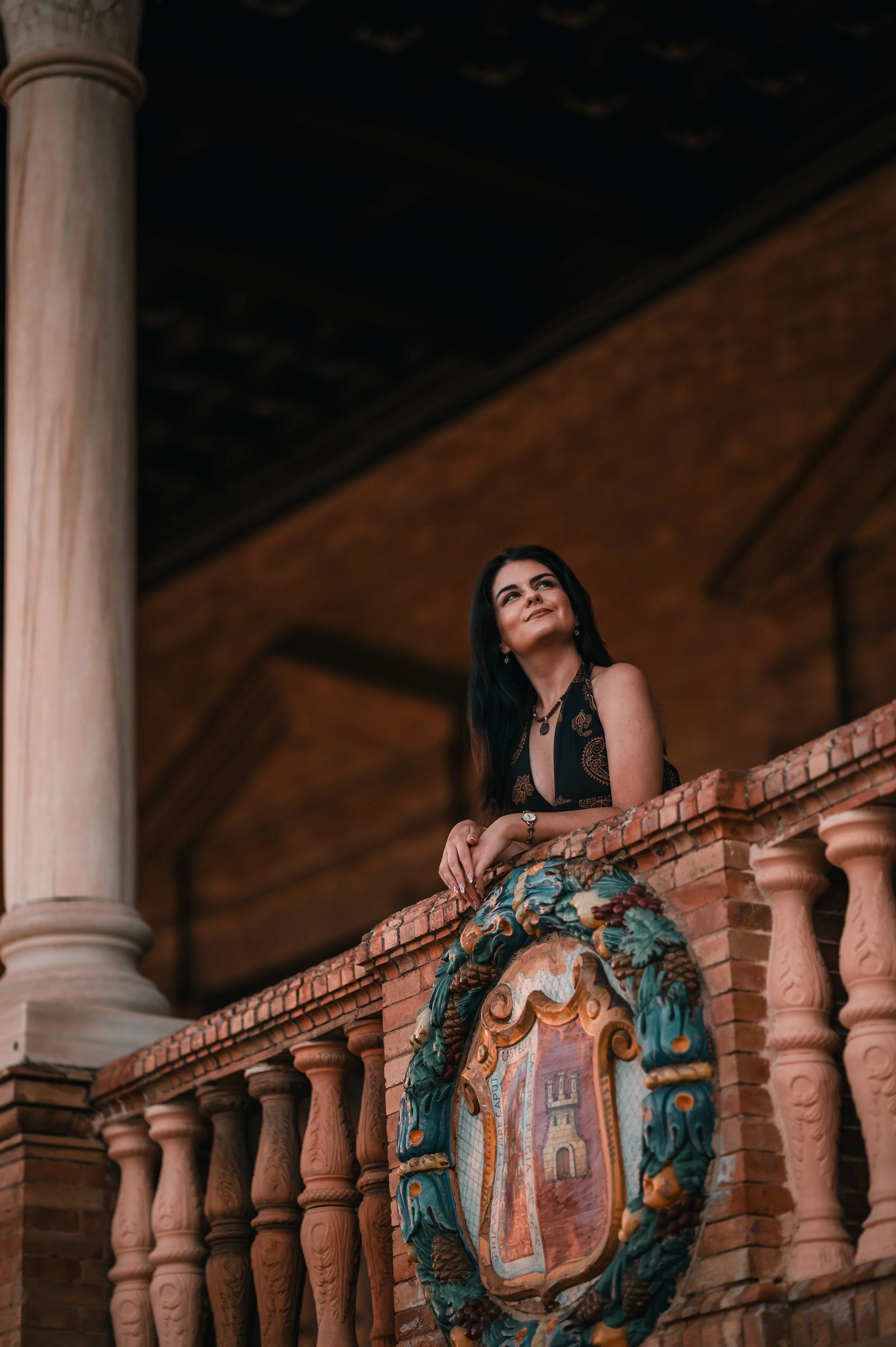 A woman with dark hair stands on a balcony overlooking a historic building, with ornate brick architecture and a colorful crest or shield on the facade.