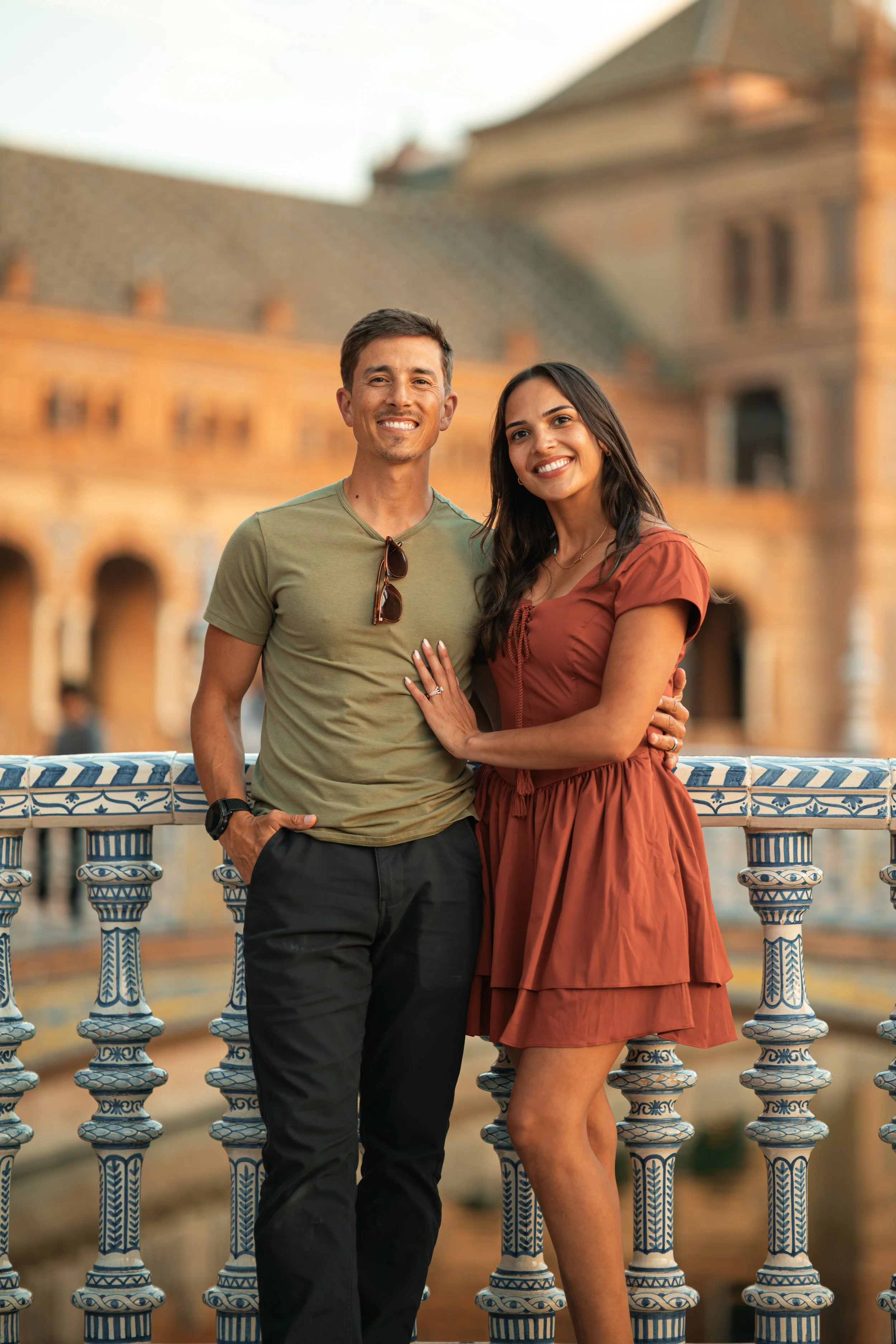 A smiling man and woman standing close together on a bridge with a decorative blue and white railing, with historic European buildings in the background.