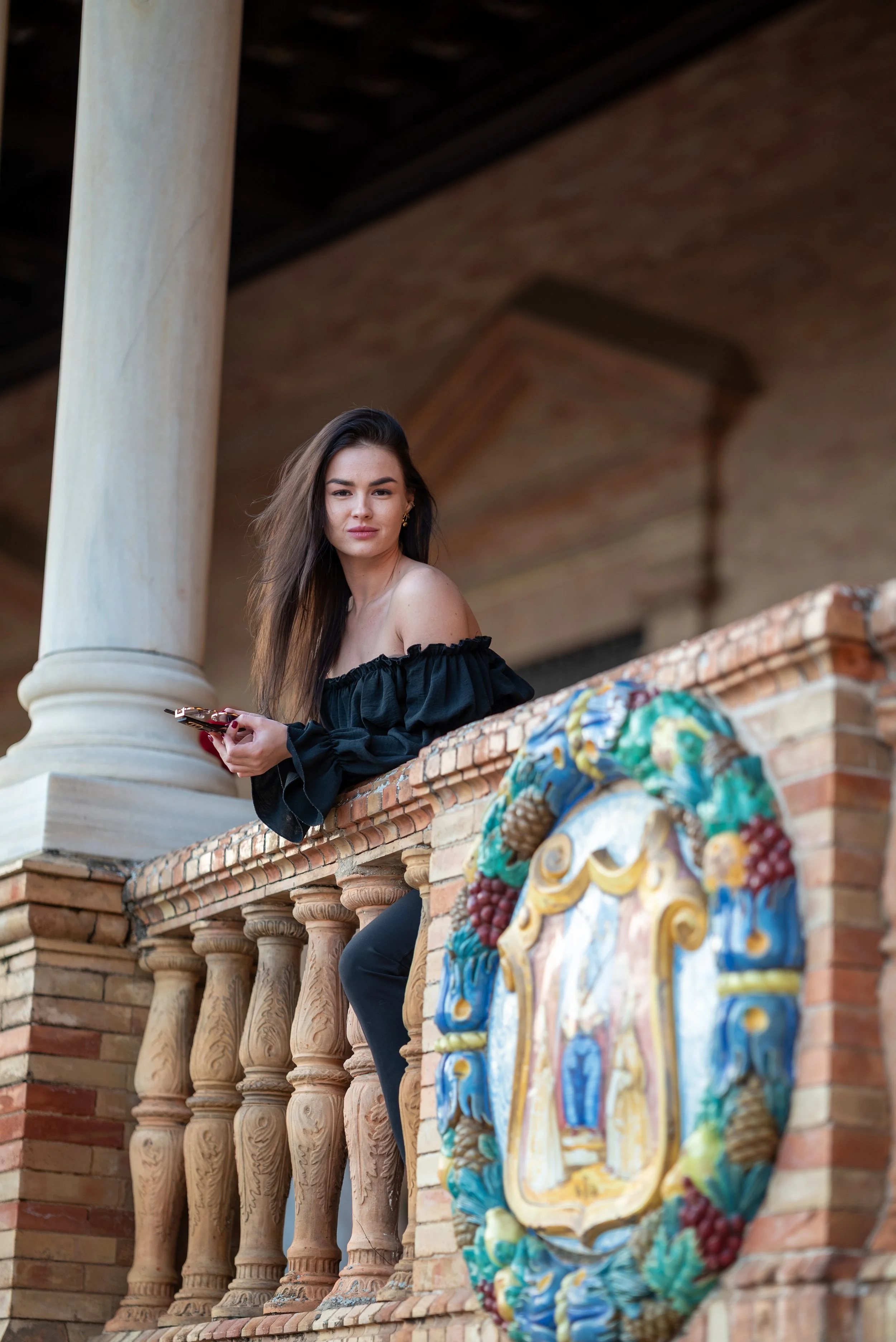 A young woman with long dark hair wearing an off-the-shoulder black top, leaning on an ornate decorative railing with detailed colorful crest, looking at the camera, on a balcony of a historical building with brick and stone architecture.