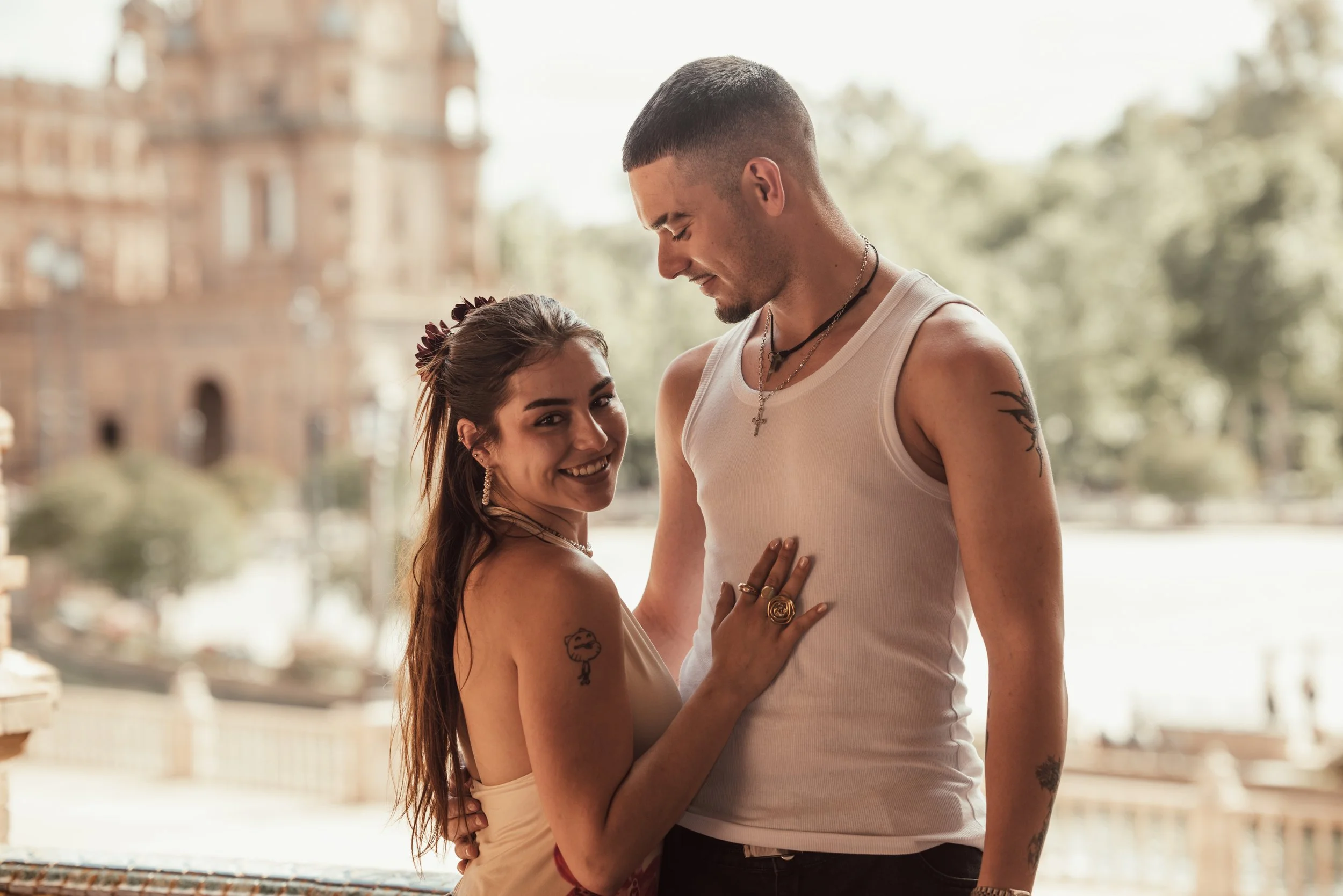 A smiling woman and a man with tattoos share an intimate moment outdoors, with a historic building in the background.
