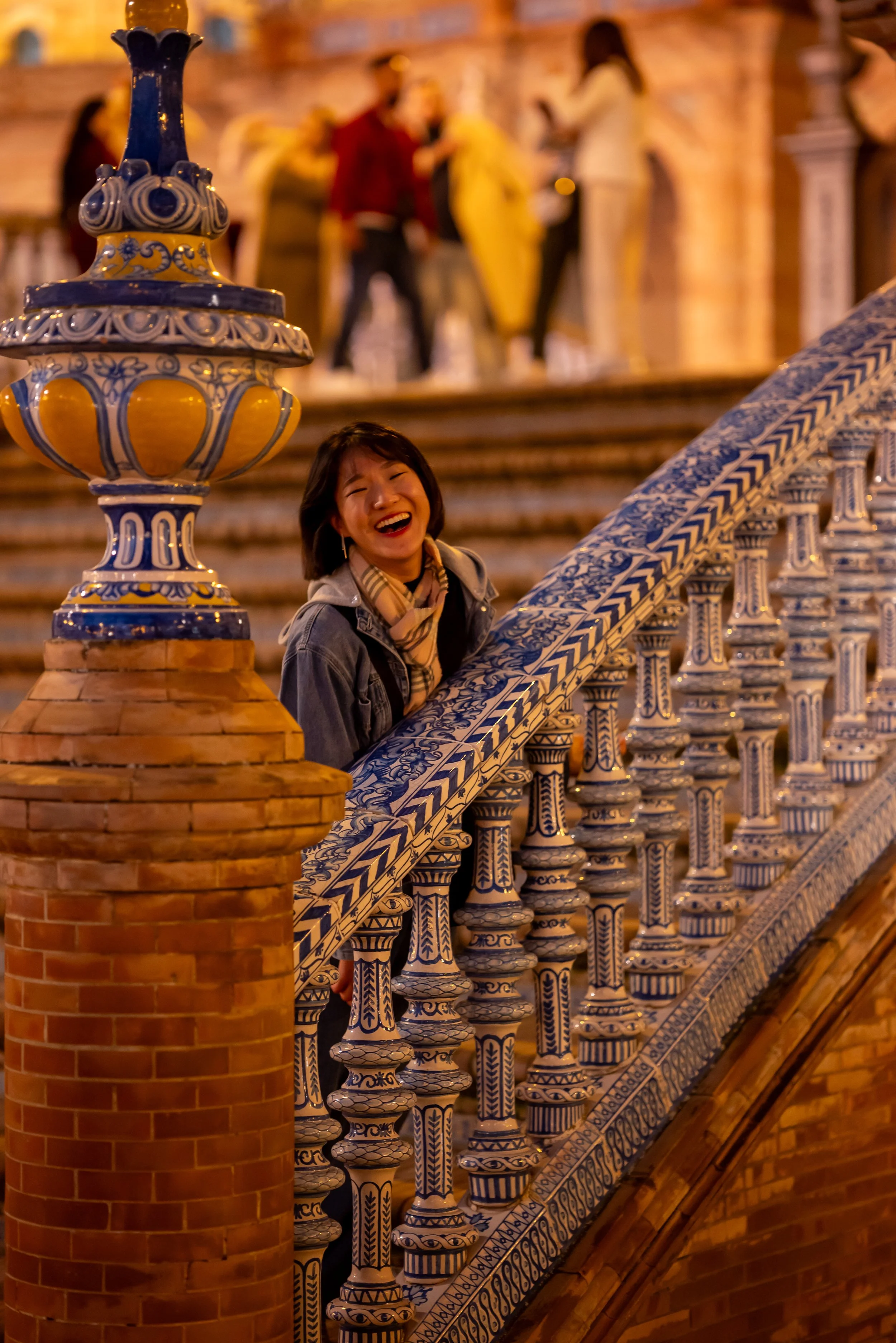 A smiling woman standing on a decorated staircase with blue and white tile designs, near a brick pillar, with blurred figures in colorful clothing in the background.