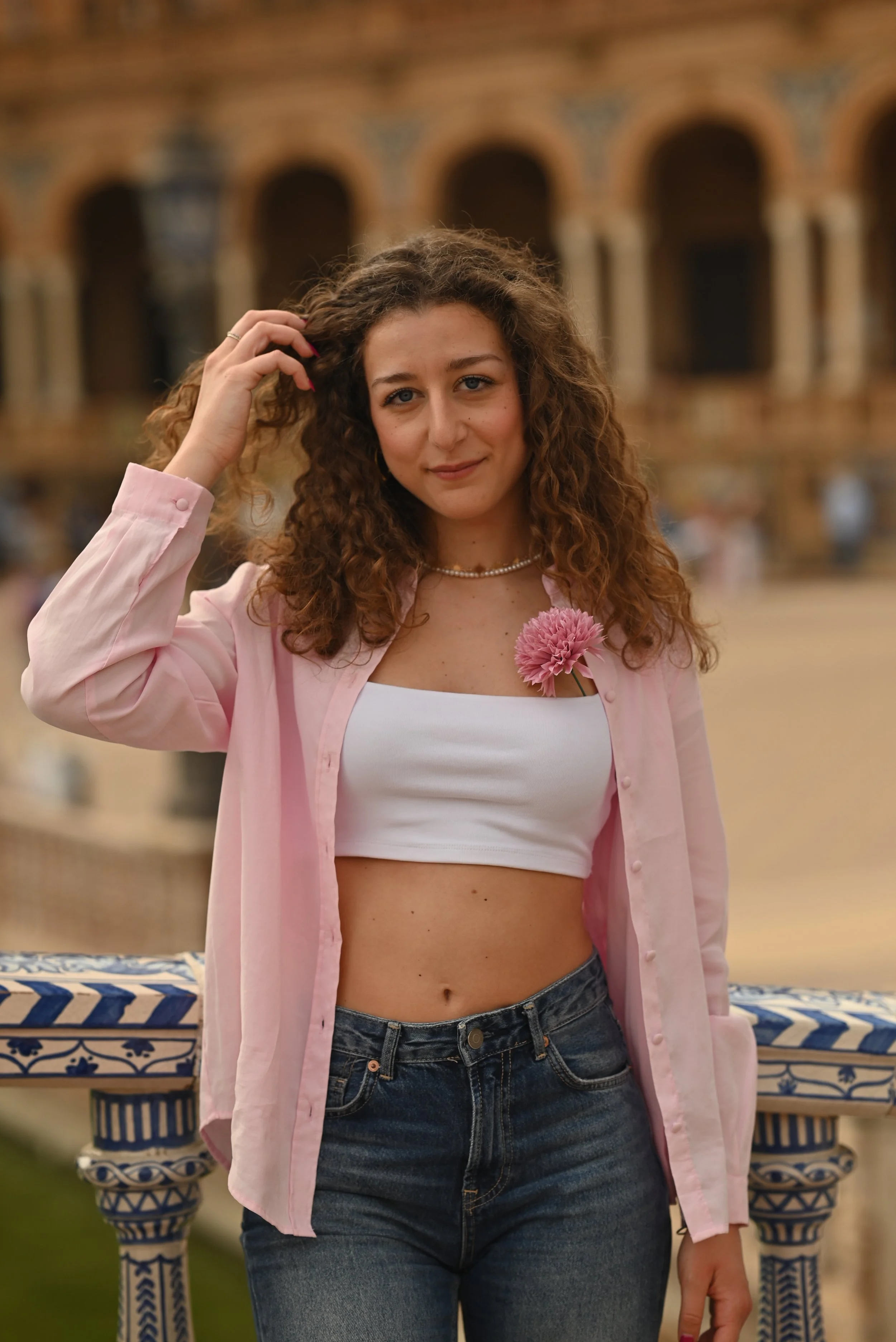 A young woman with curly brown hair and blue eyes, wearing a pink shirt over a white crop top, and jeans, holding a pink flower, standing outdoors in front of a terracotta building with arches.
