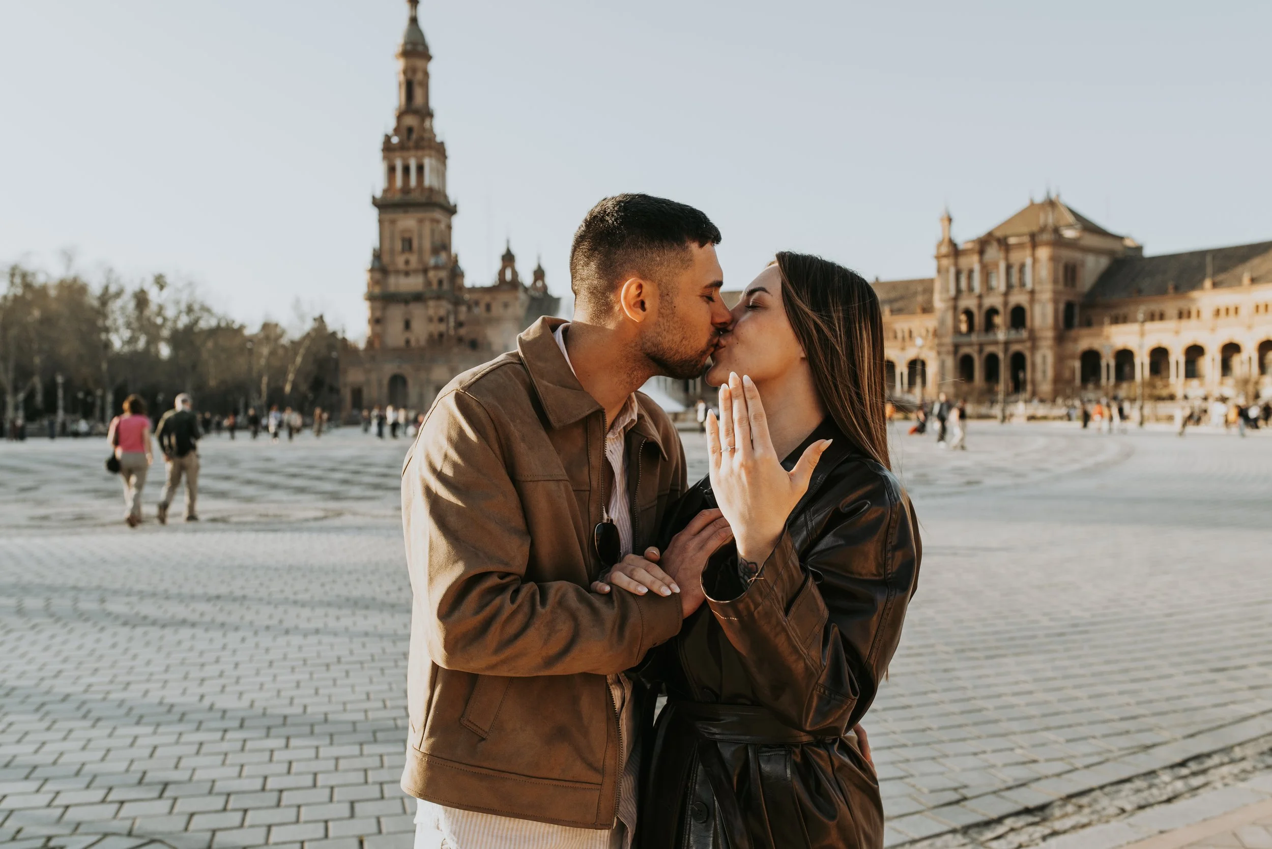 A couple is kissing in a public square with historic buildings and a tower in the background.