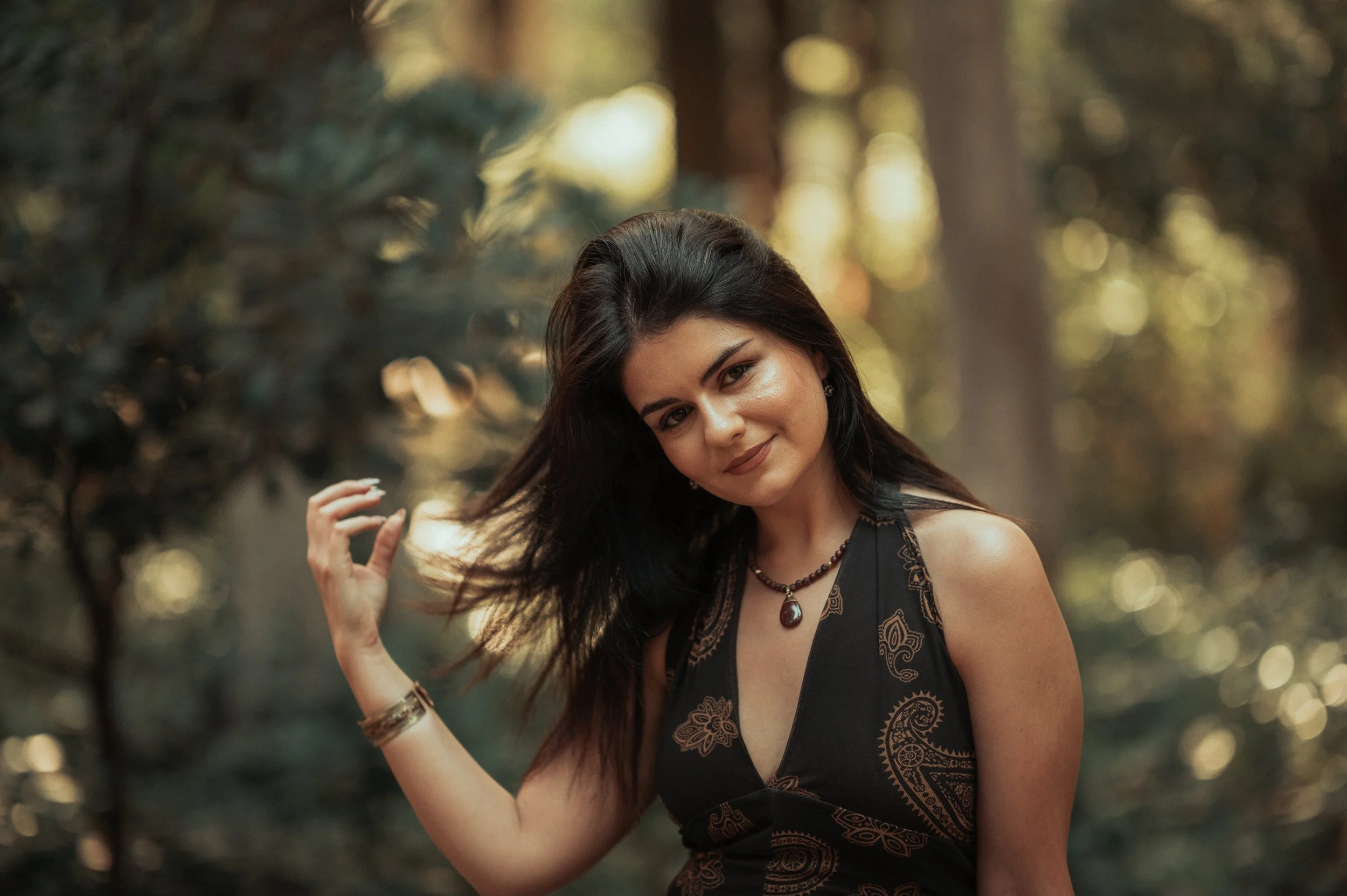 A young woman with long dark hair smiling and looking at the camera, outdoors in a wooded area during sunset, wearing a black dress with gold embroidery and jewelry.