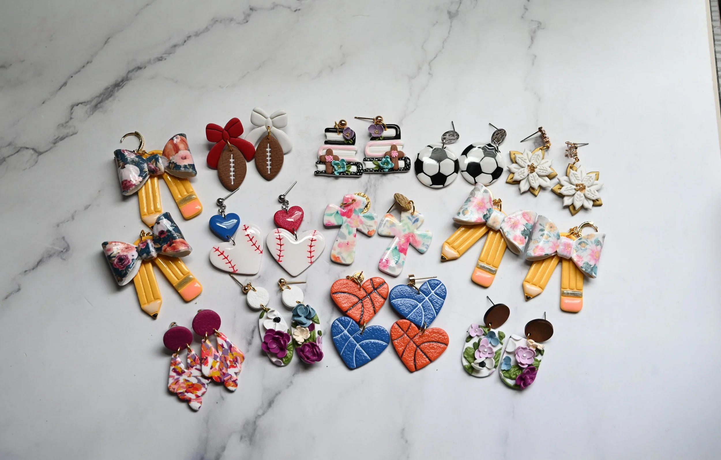 Collection of colorful earrings shaped like bows, hearts, flowers, sports balls, and abstract designs arranged on a white marble surface.