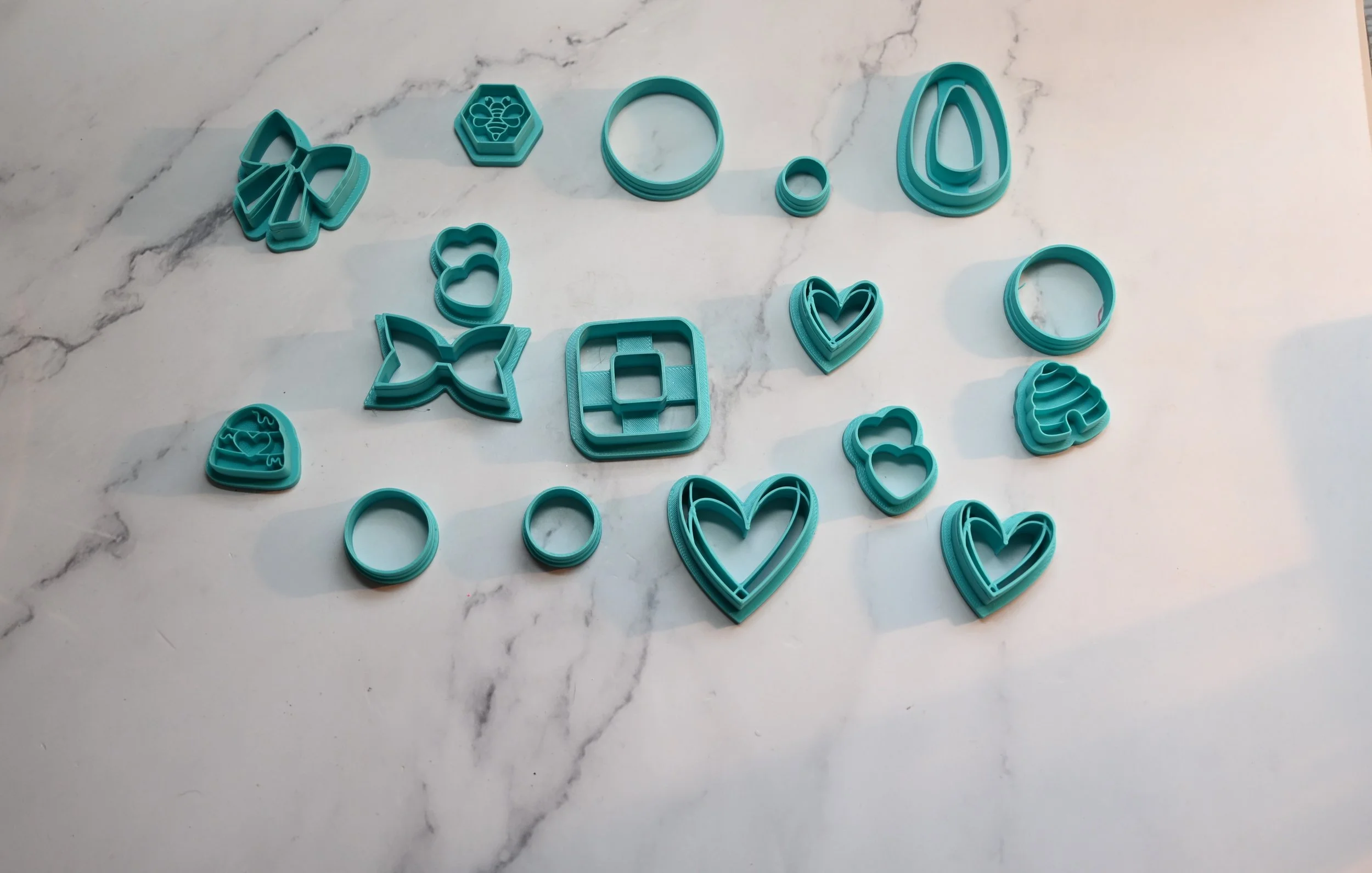 Set of teal-colored cookie cutters in various shapes, including hearts, flowers, butterflies, and geometric forms, arranged on a white marble surface.