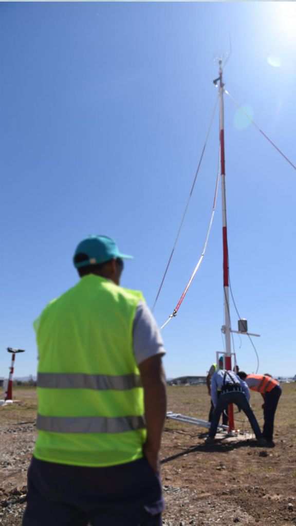 People working on setting up a tall weather mast or antenna outdoors under a clear blue sky.