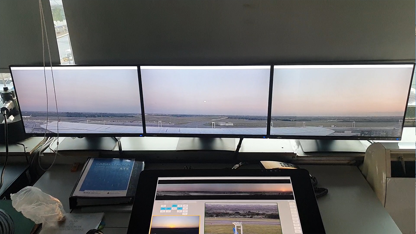 Three computer monitors showing an airport runway and sunset view, with a control tablet, a training book, and office supplies on the desk.