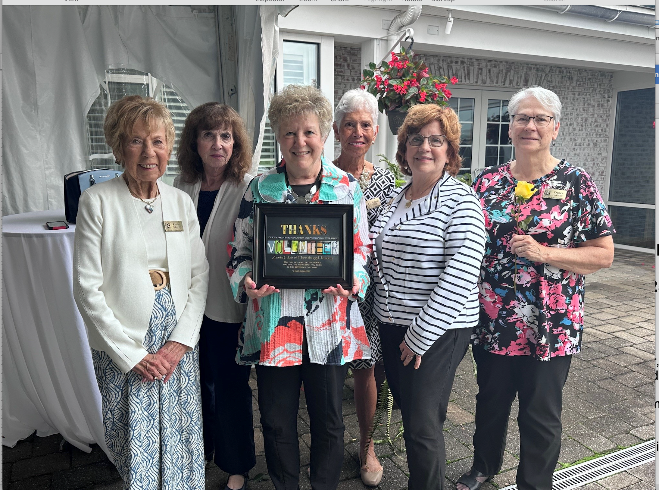 A group of six women standing together outdoors in front of a building, holding a framed sign that says 'Thanks Volunteer.' They are smiling and dressed in colorful, patterned clothing, and some are wearing name tags.
