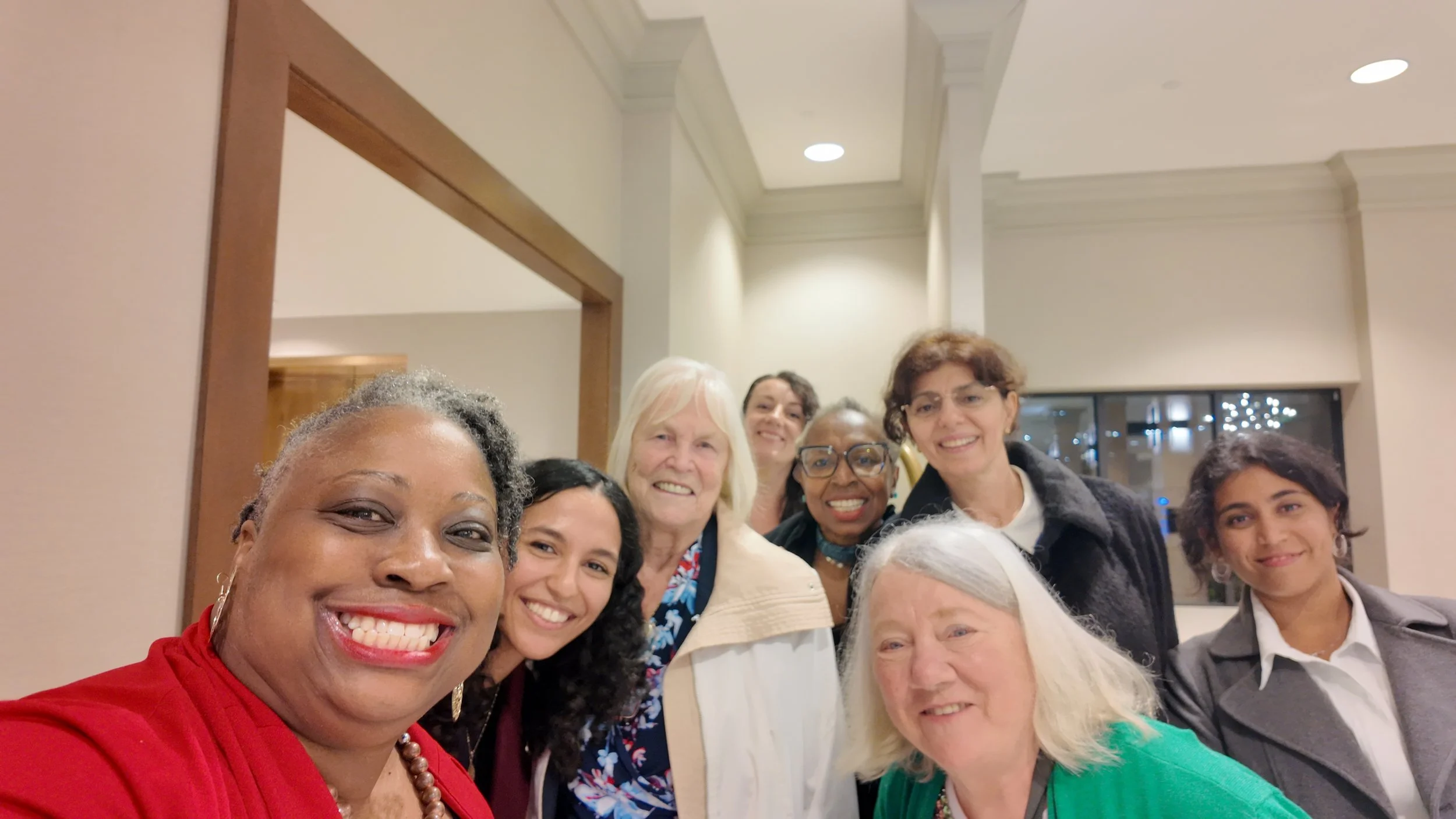 A group of diverse people smiling and posing for a photo indoors, with a beige wall and window in the background.