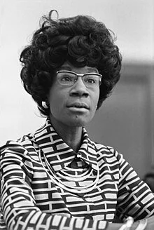Black-and-white photo of an African American woman with glasses and a large, curly hairstyle, wearing a patterned blouse and a necklace.