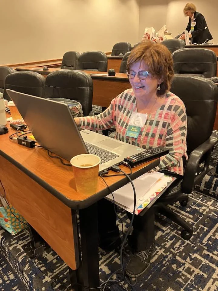 A woman with glasses sitting at a conference table, working on a laptop with a coffee cup in front of her, in a conference room with empty seats. A person is standing in the background.