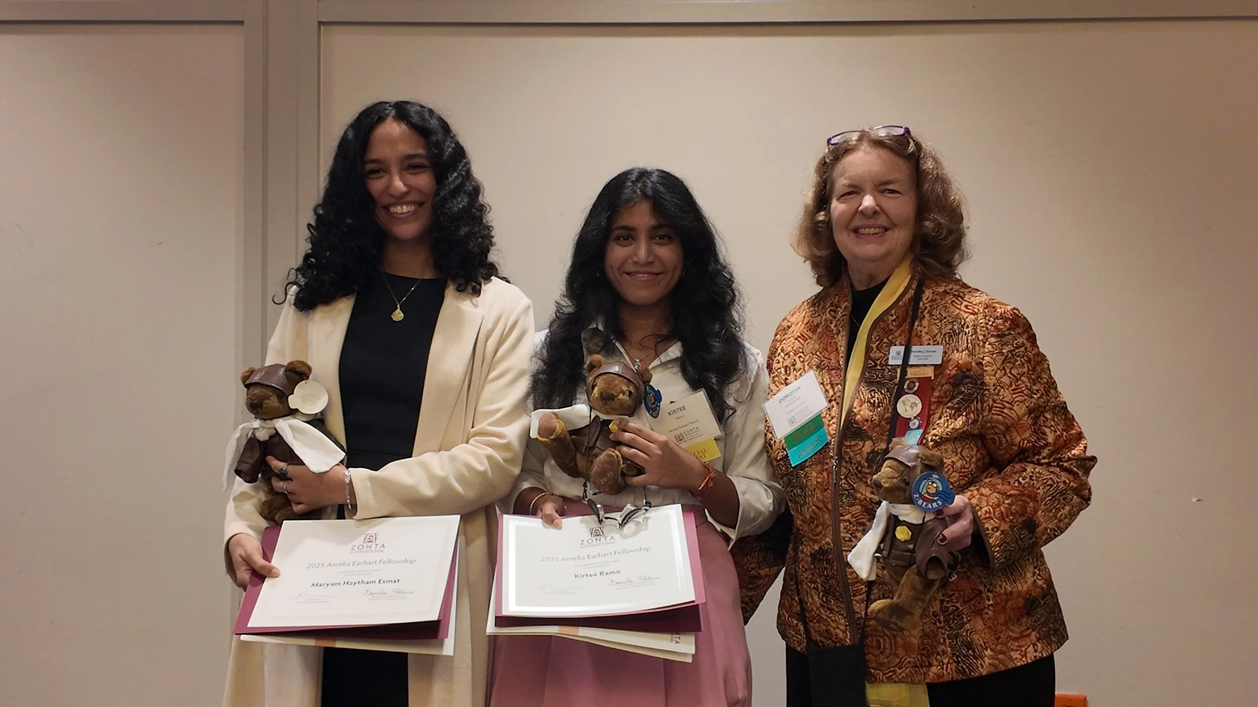 Three women standing together, each holding a teddy bear and certification plaques, smiling at a conference or award event, with woman on the far right wearing glasses resting on her head and dressed in a orange patterned blazer.