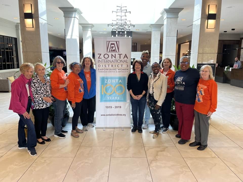 Group of women standing around a Zonta International District 3 sign at a conference, celebrating Zonta International's 100th anniversary, with modern hotel lobby background.