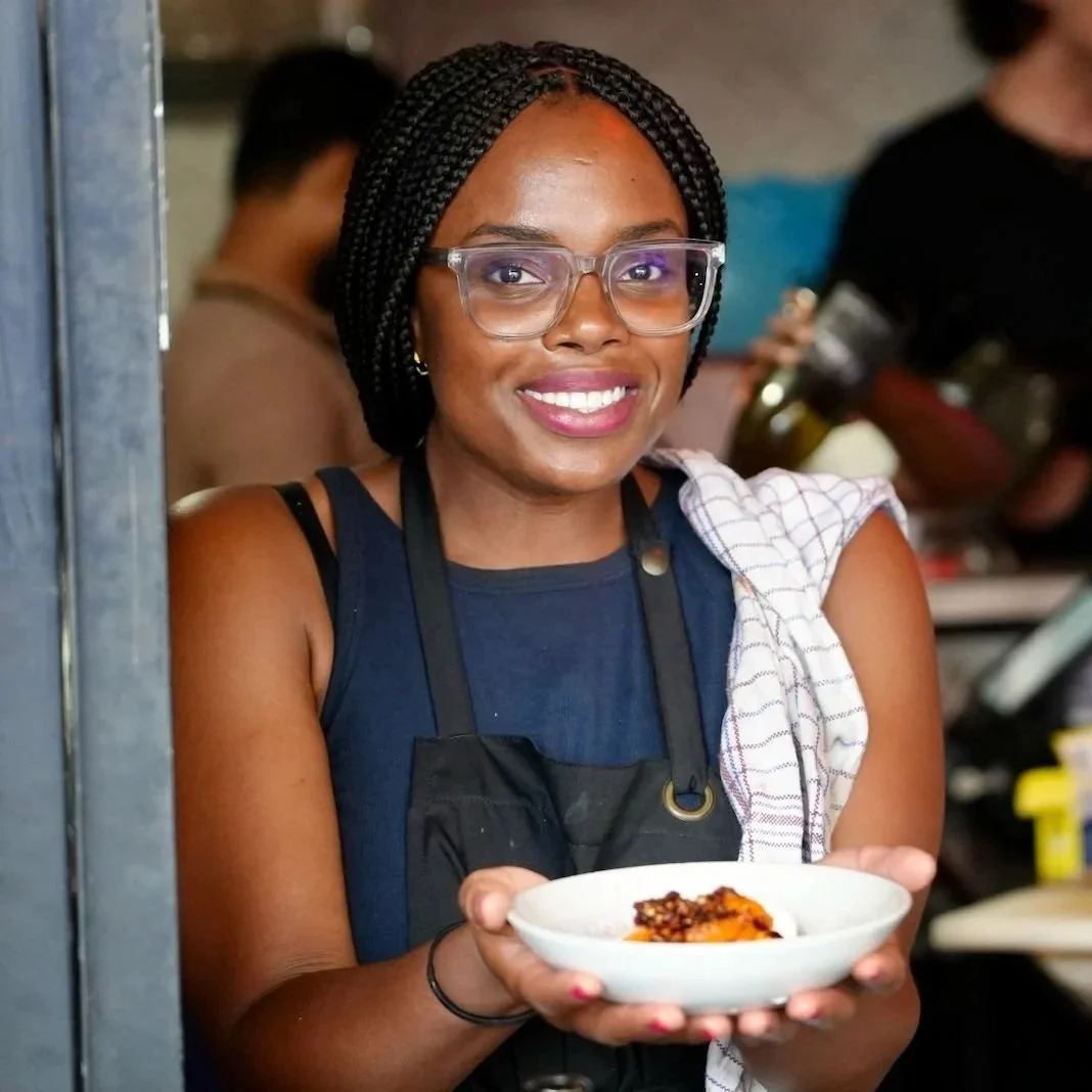A woman with glasses smiling while holding a bowl of food in a busy kitchen or restaurant setting.