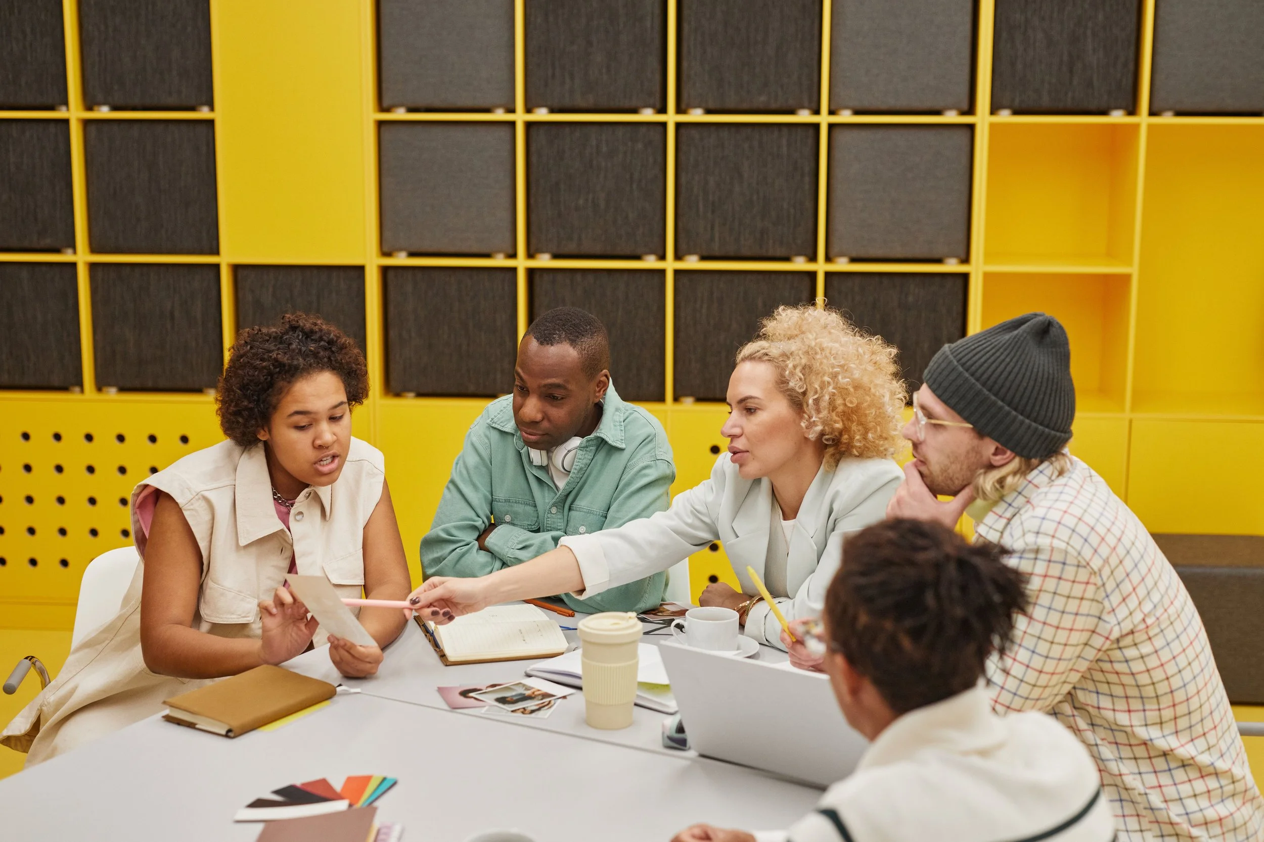 A group of five people sitting around a table in a modern room with bright yellow wall storage is engaged in a discussion. They have notebooks, photos, color swatches, and drinks on the table.