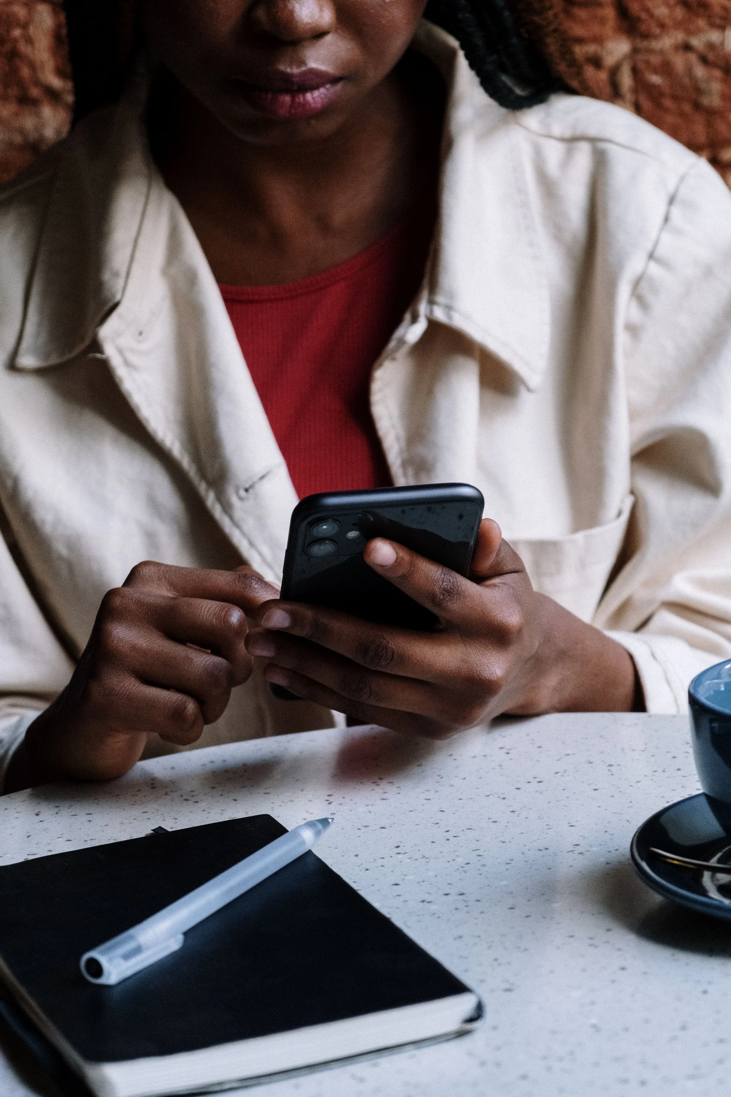 A person with dark skin and braided hair wearing a white jacket and red shirt, sitting at a table and looking at a smartphone.