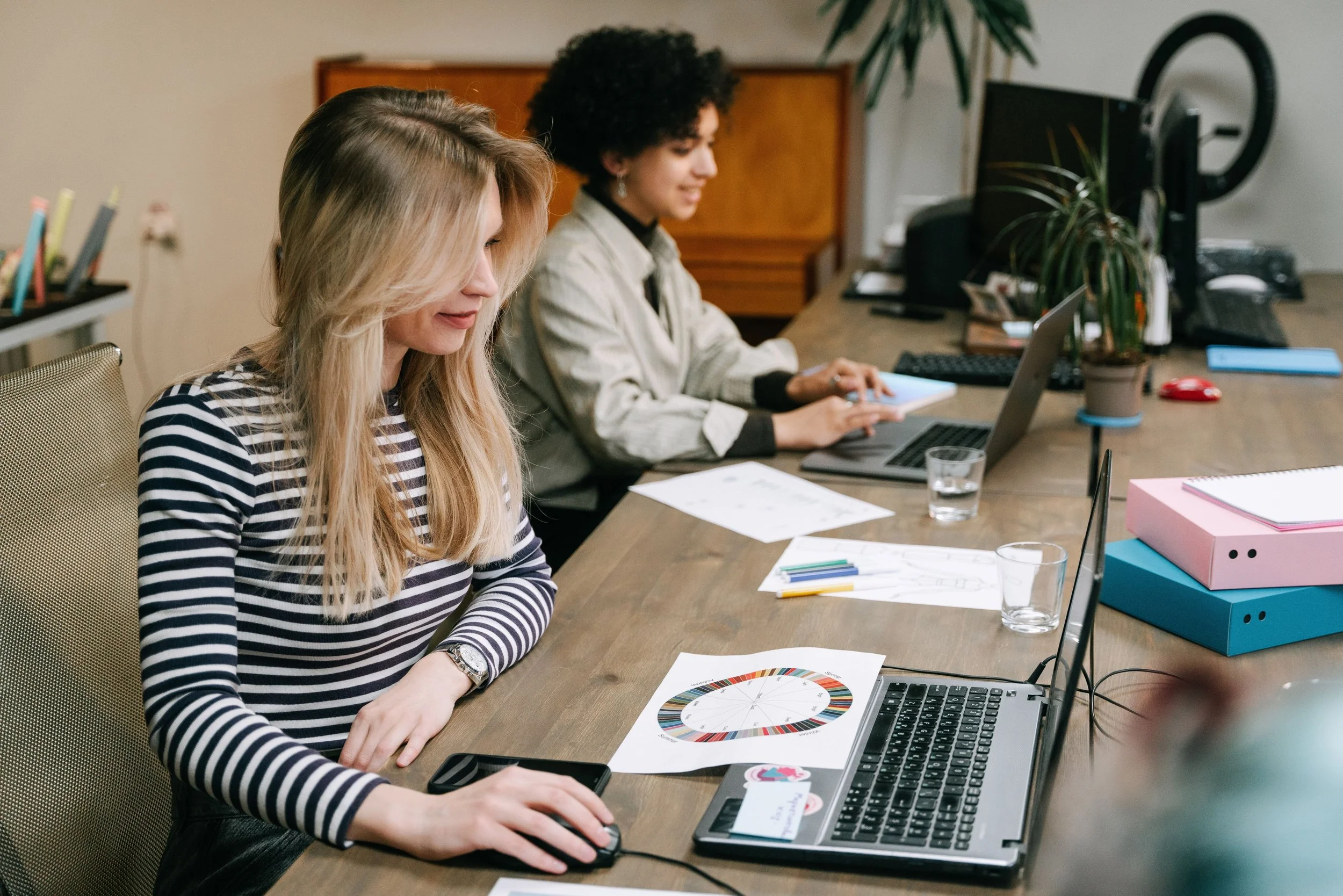Two women working at a desk with laptops, documents, and office supplies, including a color wheel, in a professional office environment.